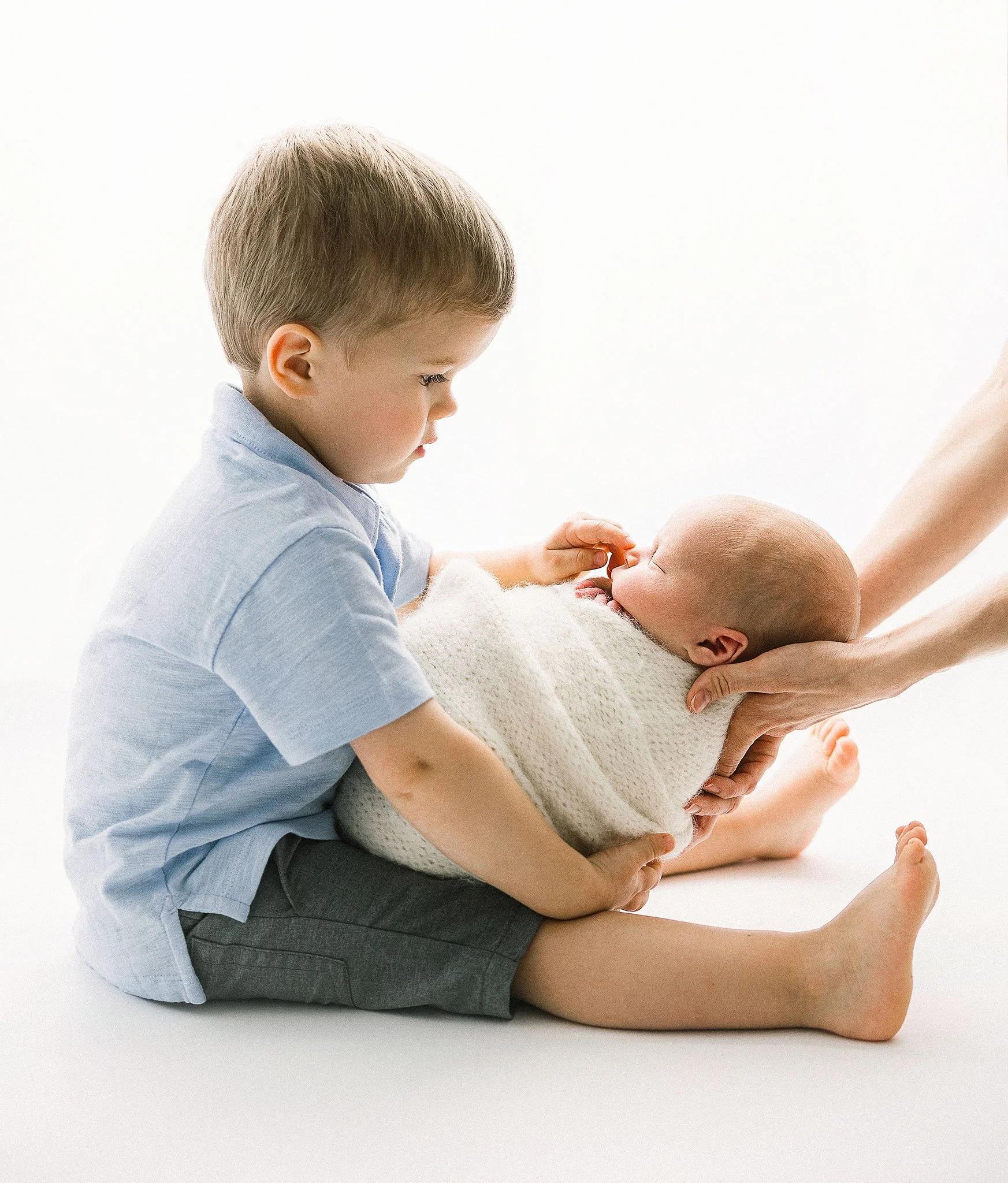 A young boy touching a newborn baby's nose while the baby is held in an adult's hands, on a white background.