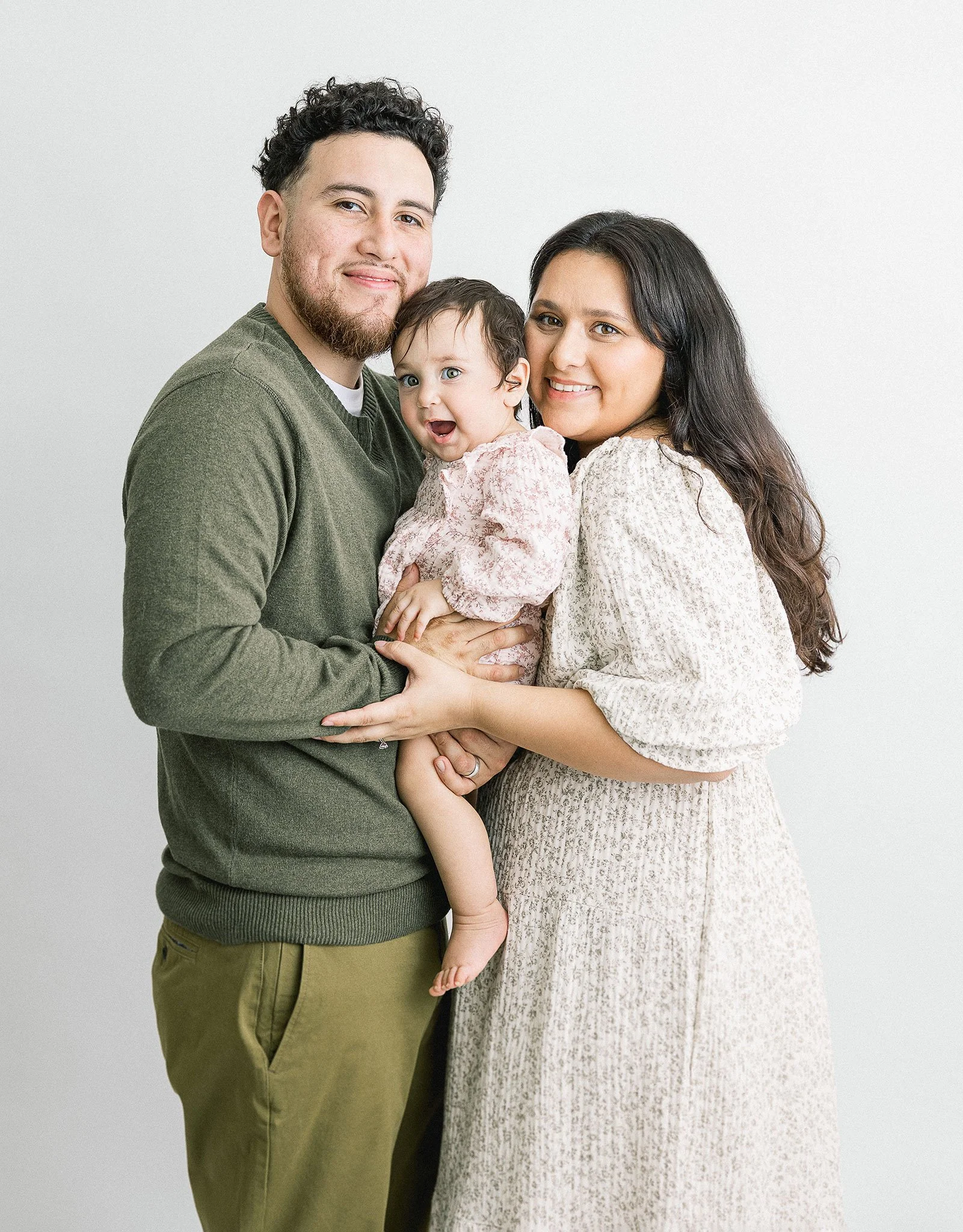 A man and a woman, both smiling, holding a baby between them. The man is on the left wearing a green sweater and khaki pants, the woman is on the right in a light-colored floral dress, and the baby is dressed in a light pink outfit.