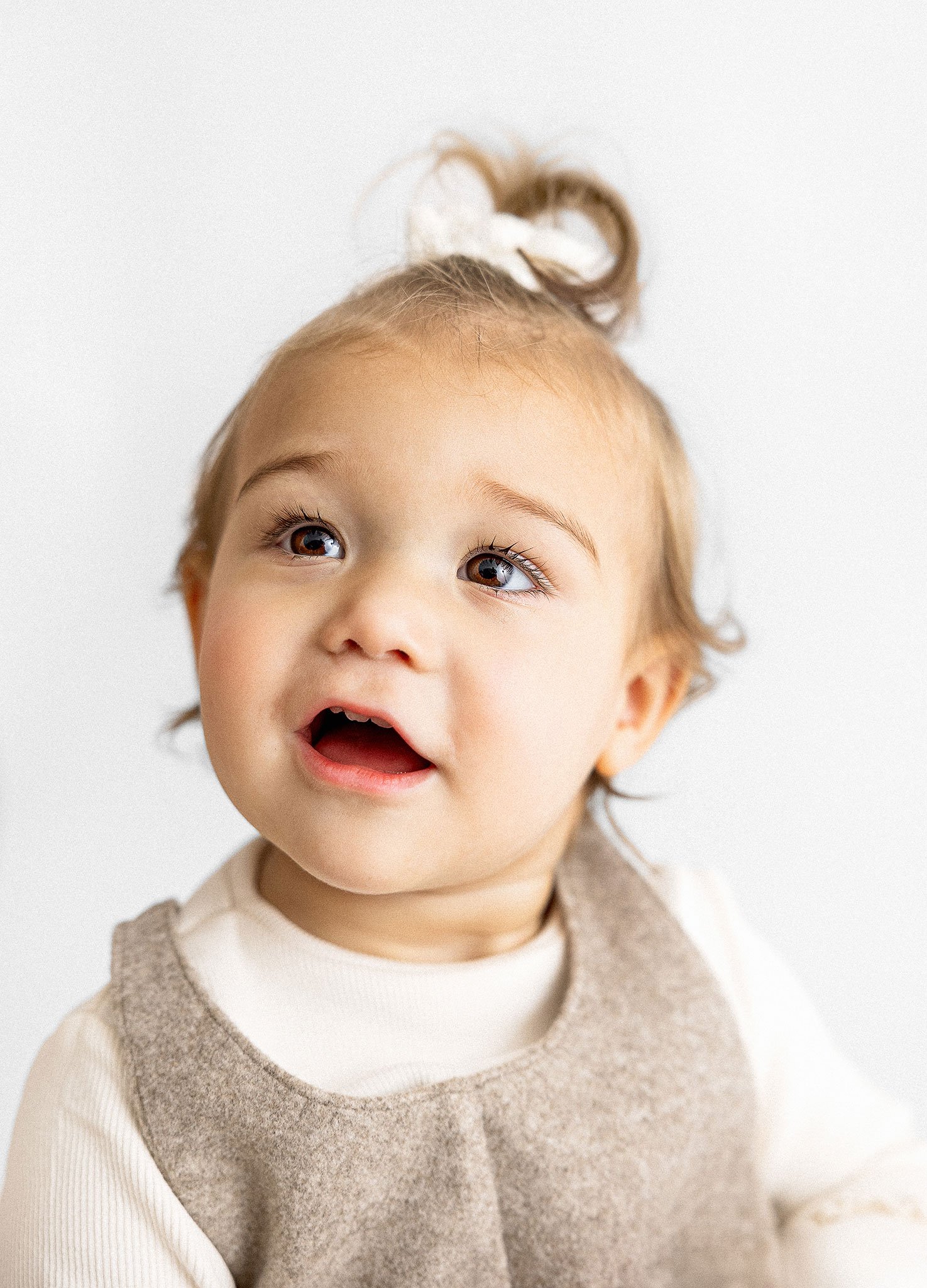Close-up of a toddler looking up with a curious expression, wearing a beige top and a white hair bow.