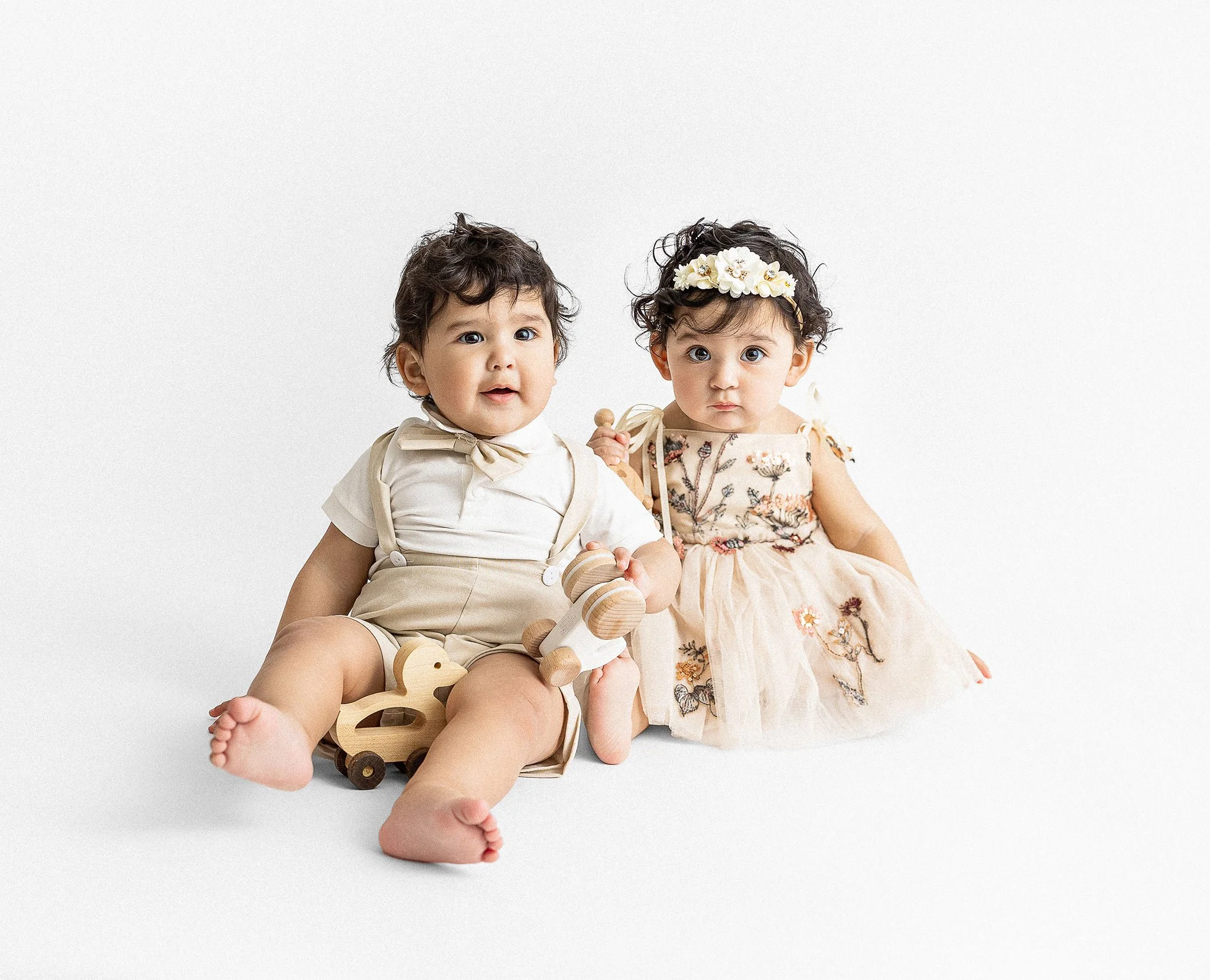 Two young children sitting together on a plain white background. The child on the left has short, curly dark hair, blue eyes, and is wearing cream-colored shorts and a shirt with beige suspenders. The child on the right has curly dark hair adorned wi