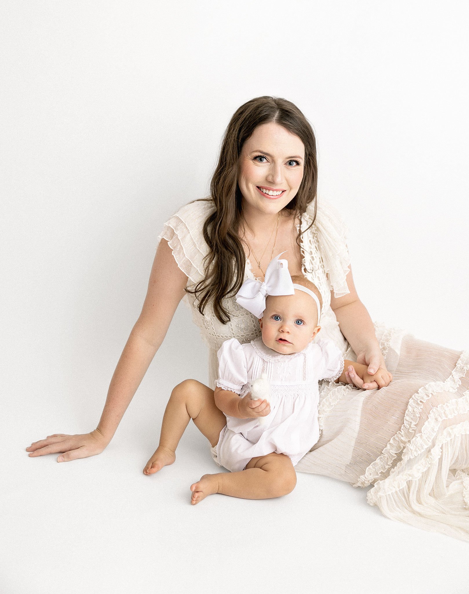 Mother posing with baby in white outfits against plain background.