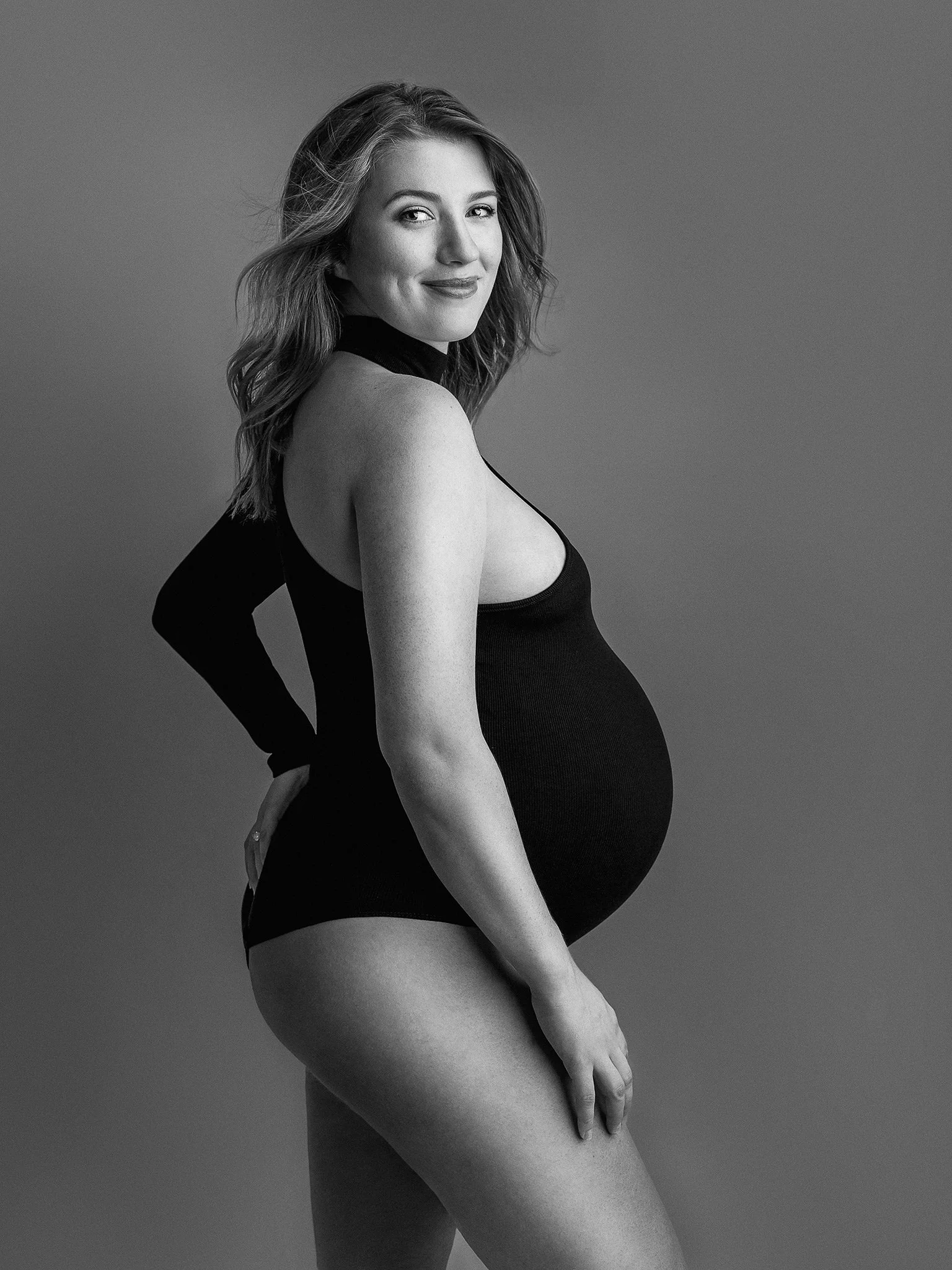 Pregnant woman in black bodysuit, side view, smiling, black and white.
