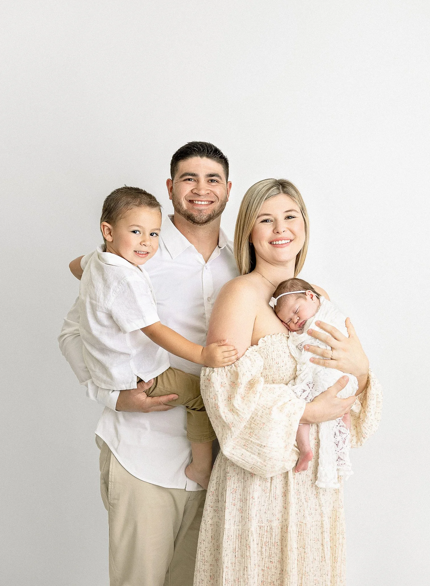 A smiling family of four, including a mother holding a newborn, father, and young son, posing against a plain white background.