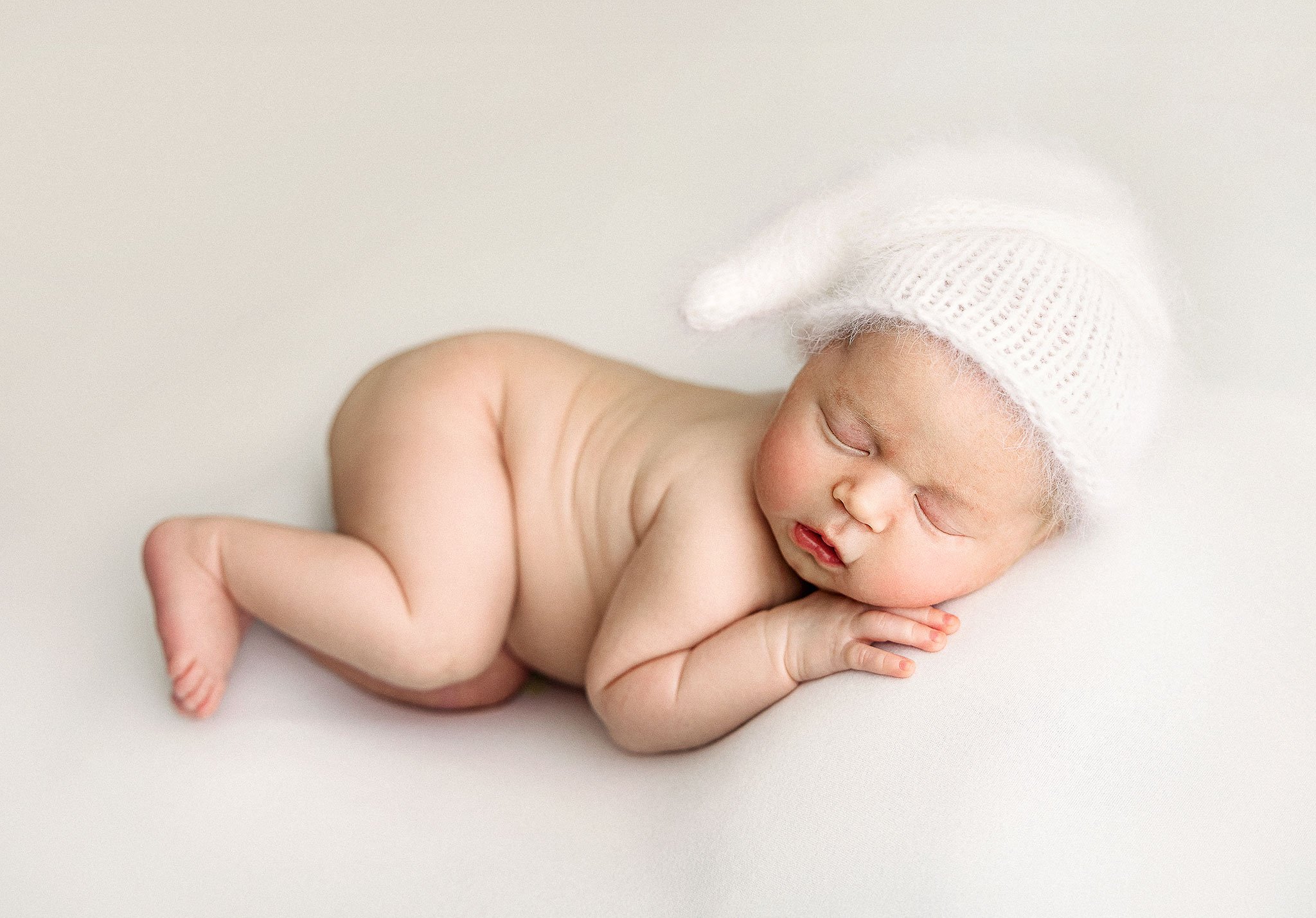Newborn baby sleeping, wearing a white knit hat, lying on a light surface.