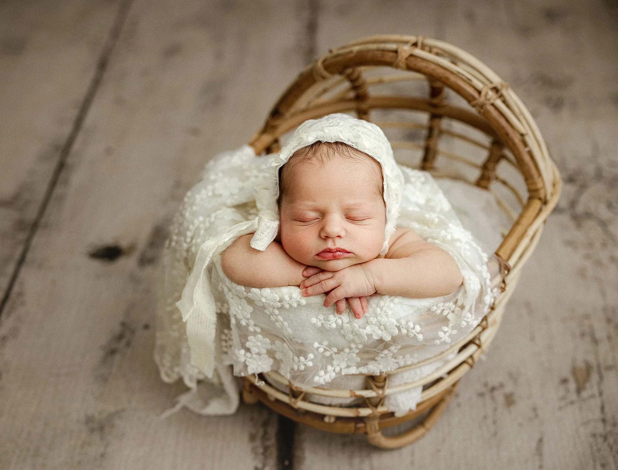 Newborn baby sleeping in a wooden basket, wrapped in a white lace blanket, wearing a matching bonnet.