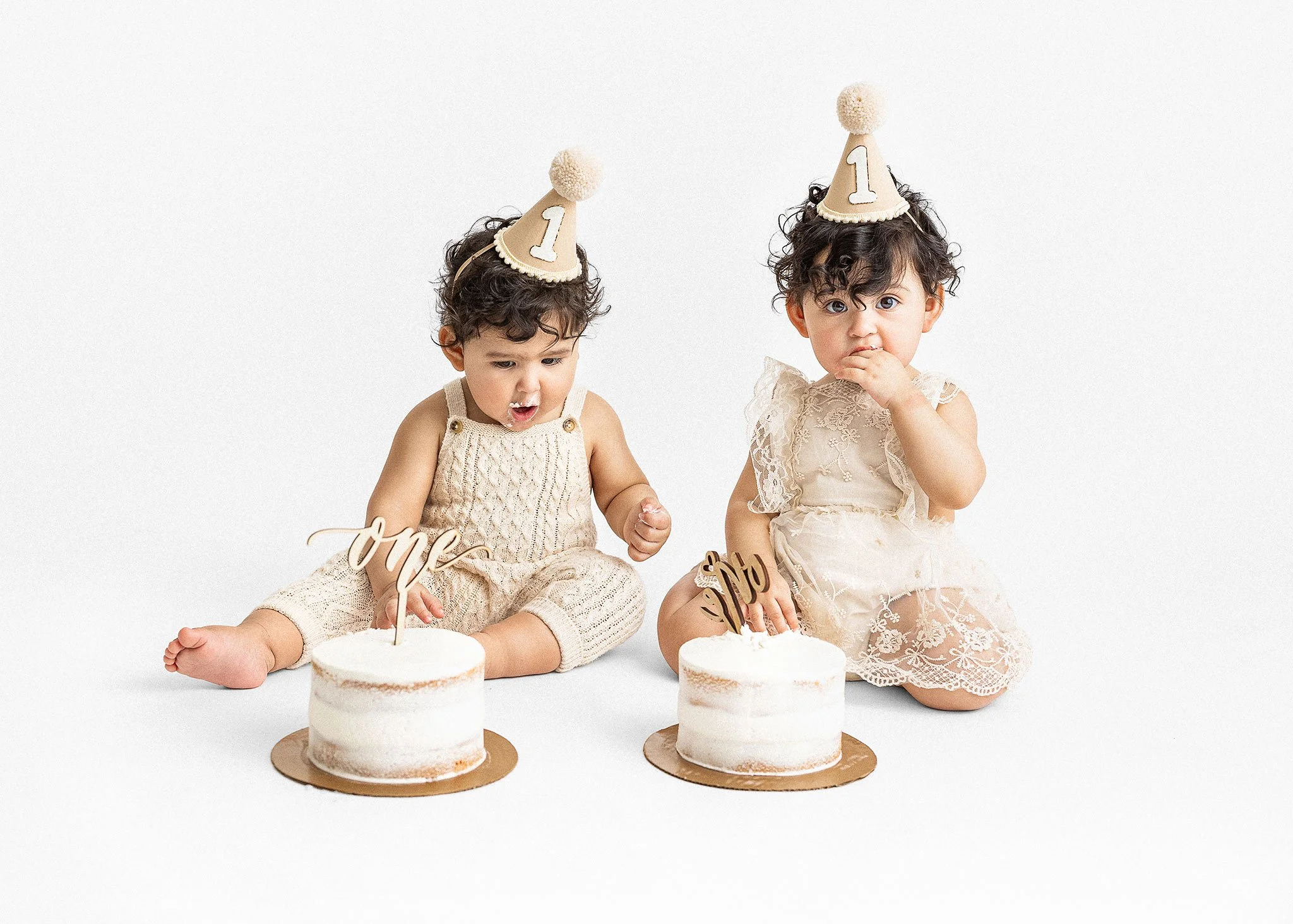 Two young children with curly hair wearing beige birthday hats, sitting in front of small cakes. The cake on the left has a topper that reads 'One', and the cake on the right has a topper that reads 'Uno'. The children are dressed in cream-colored ou