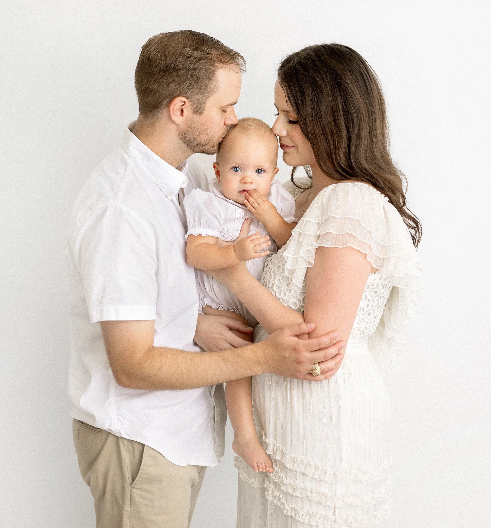 A family portrait featuring a man, woman, and baby dressed in white, standing closely together against a plain background.