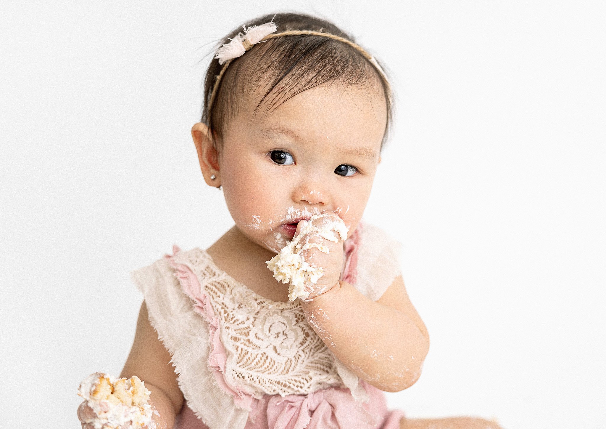 Baby with headband eating cake, wearing a lace dress, and looking at the camera.