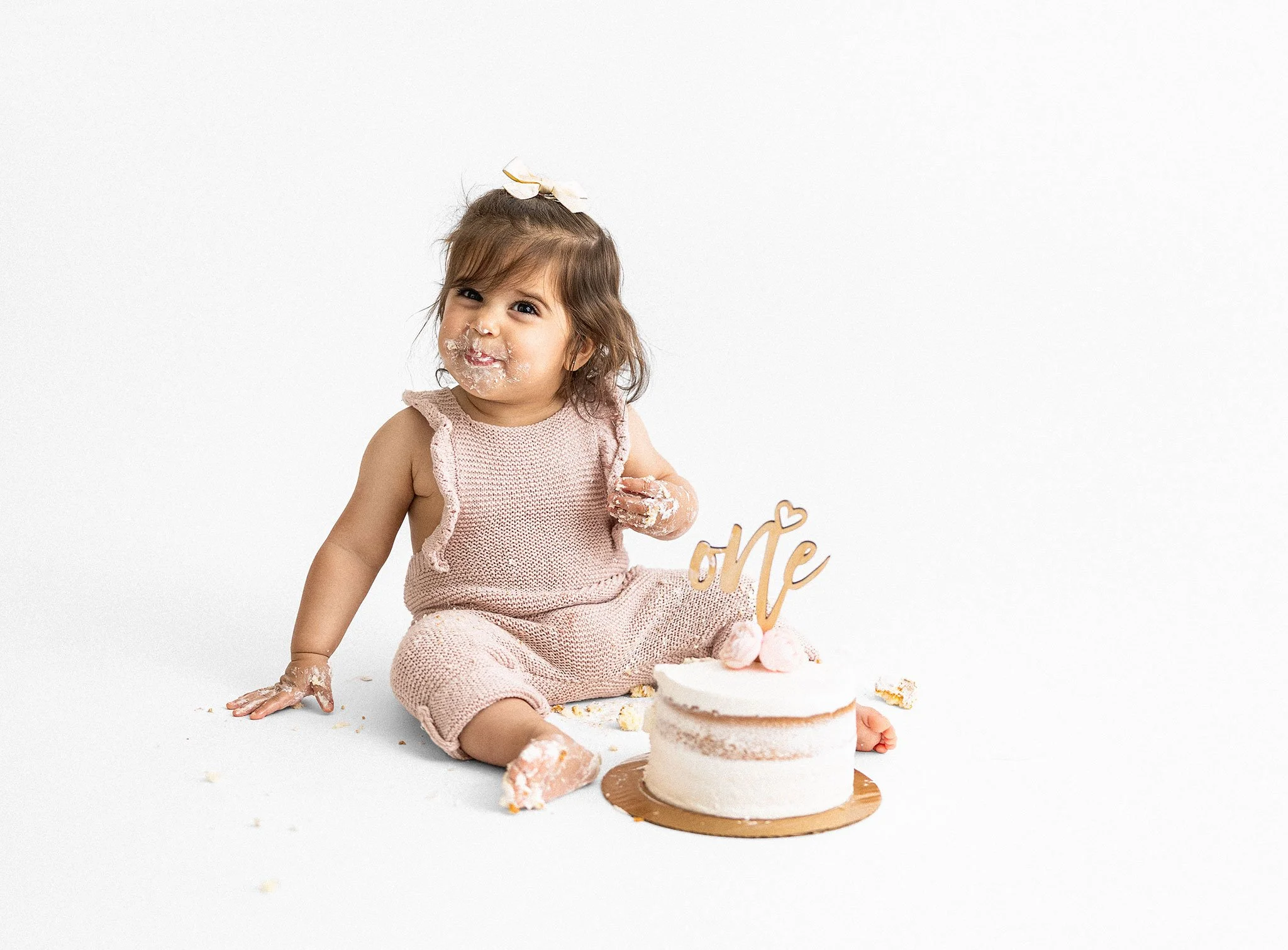 Cute baby girl with messy face and hands sitting on the white floor, next to a small white frosted cake with pink decorations and a wooden "one" cake topper for her first birthday celebration.