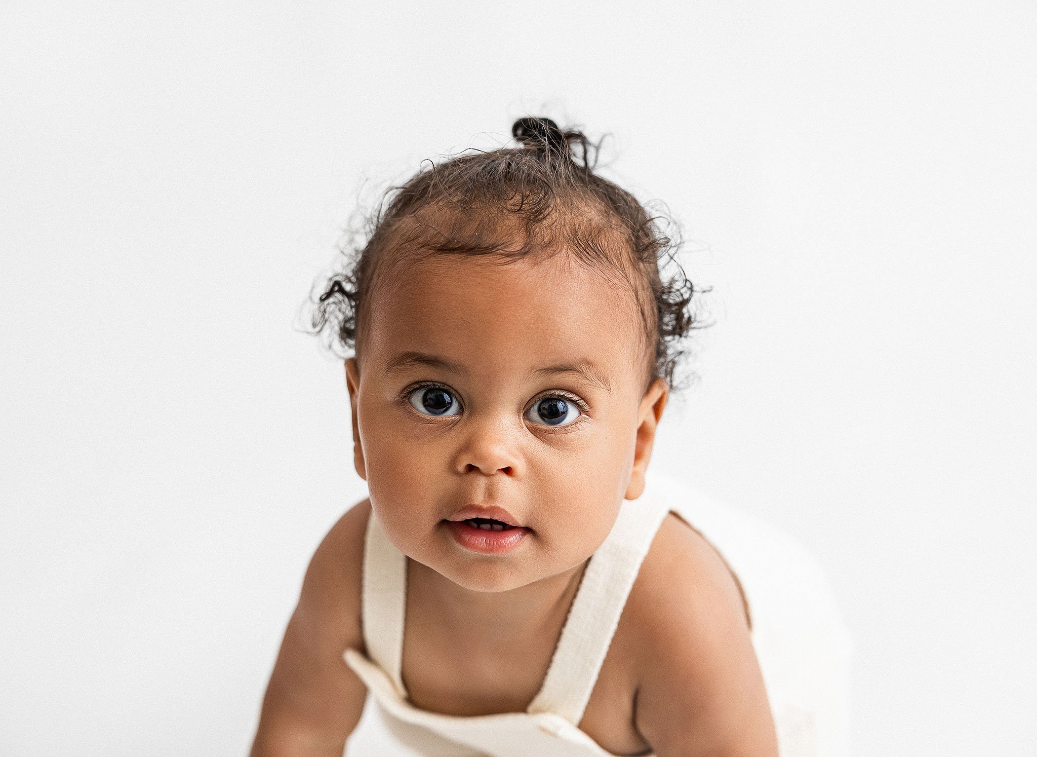 Baby with curly hair wearing a cream-colored outfit, looking at the camera against a white background.