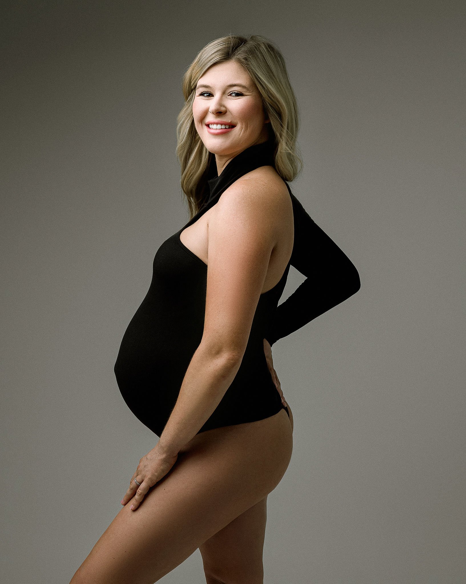 Pregnant woman in a black bodysuit posing against a gray background.
