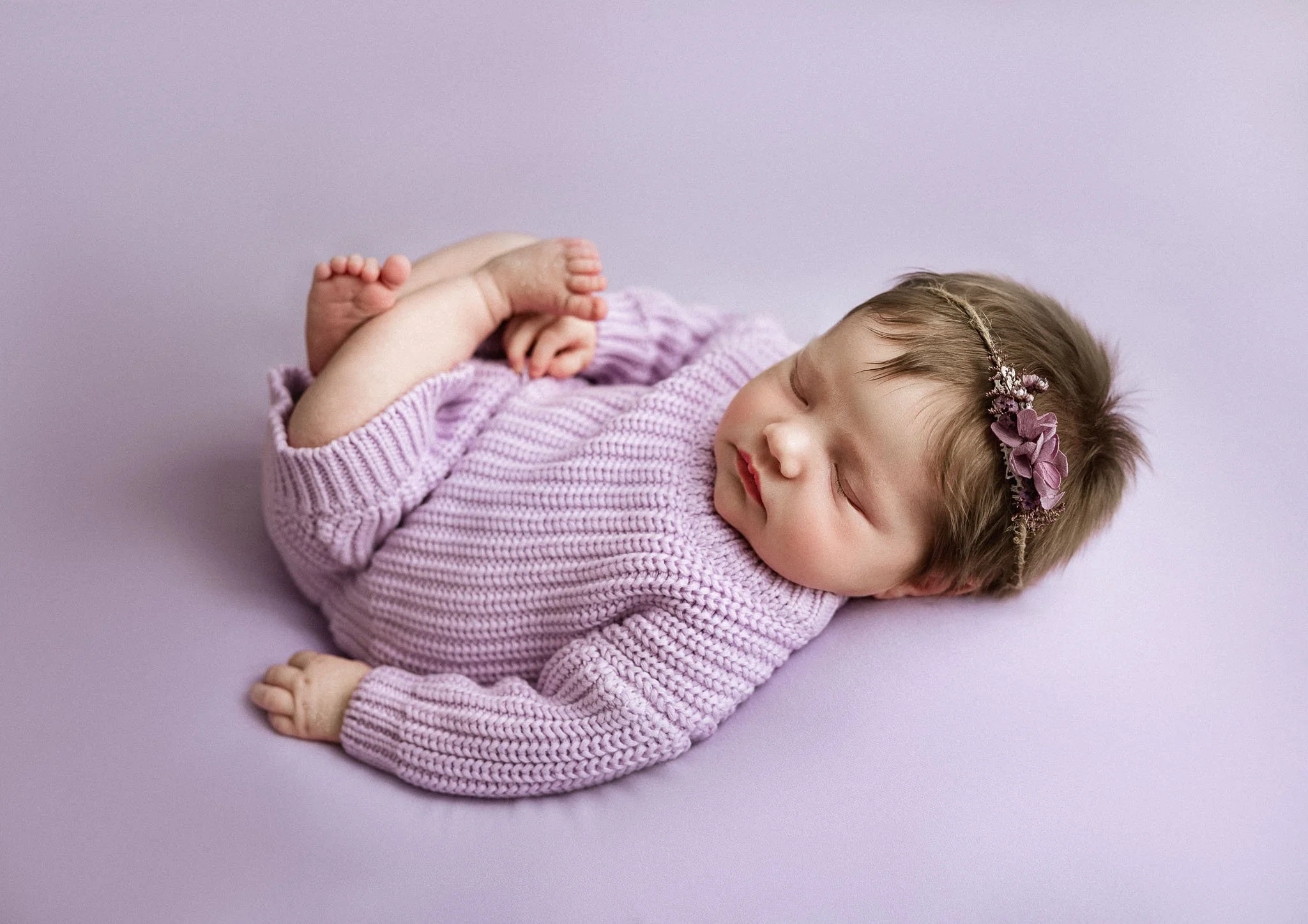 A young girl with light brown hair, wearing a pink knitted sweater and a floral headband, is sleeping on her side with her arms crossed, lying on a purple surface against a purple background.