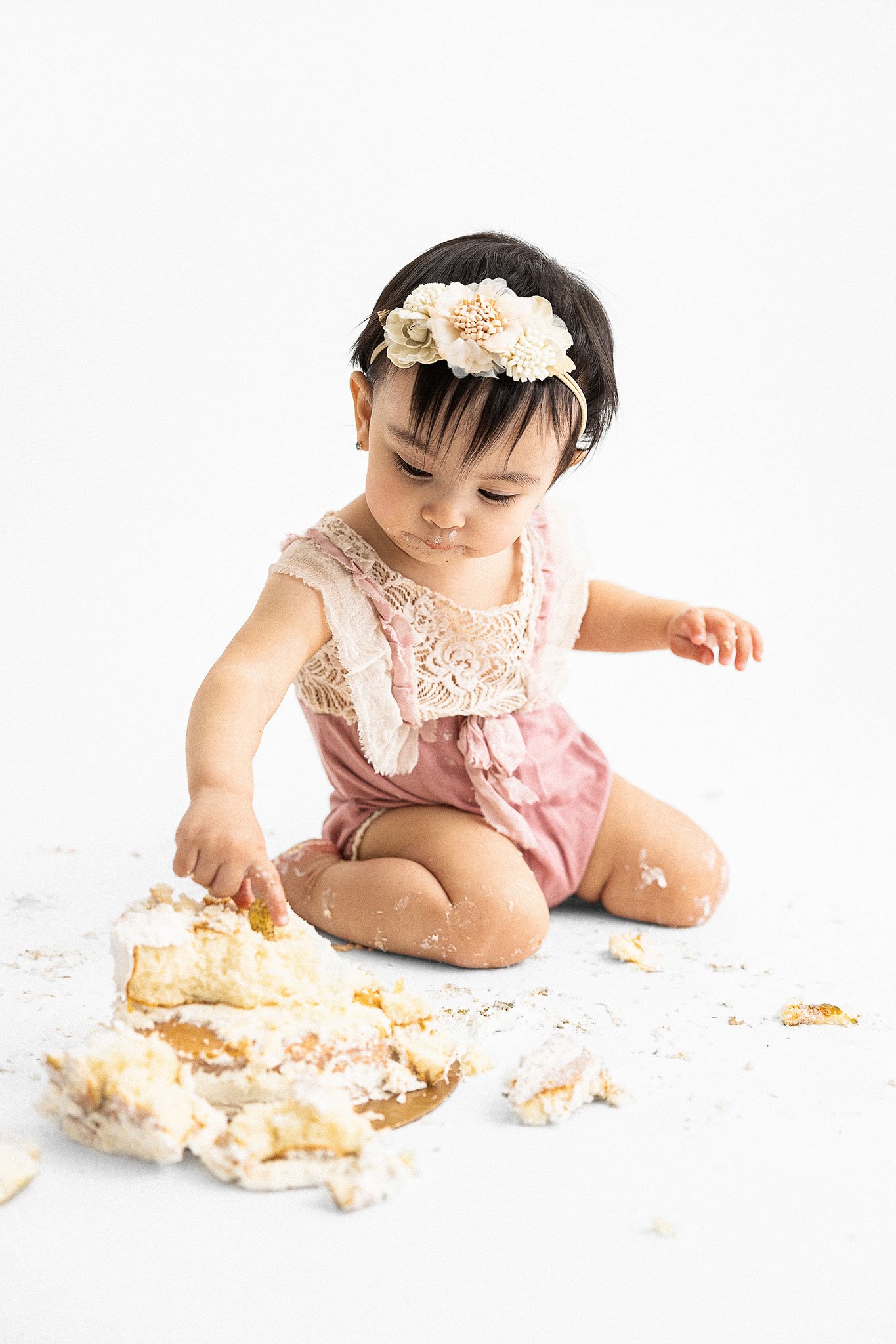 Baby in pink lace outfit playing with a smashed cake on the floor, wearing a floral headband.