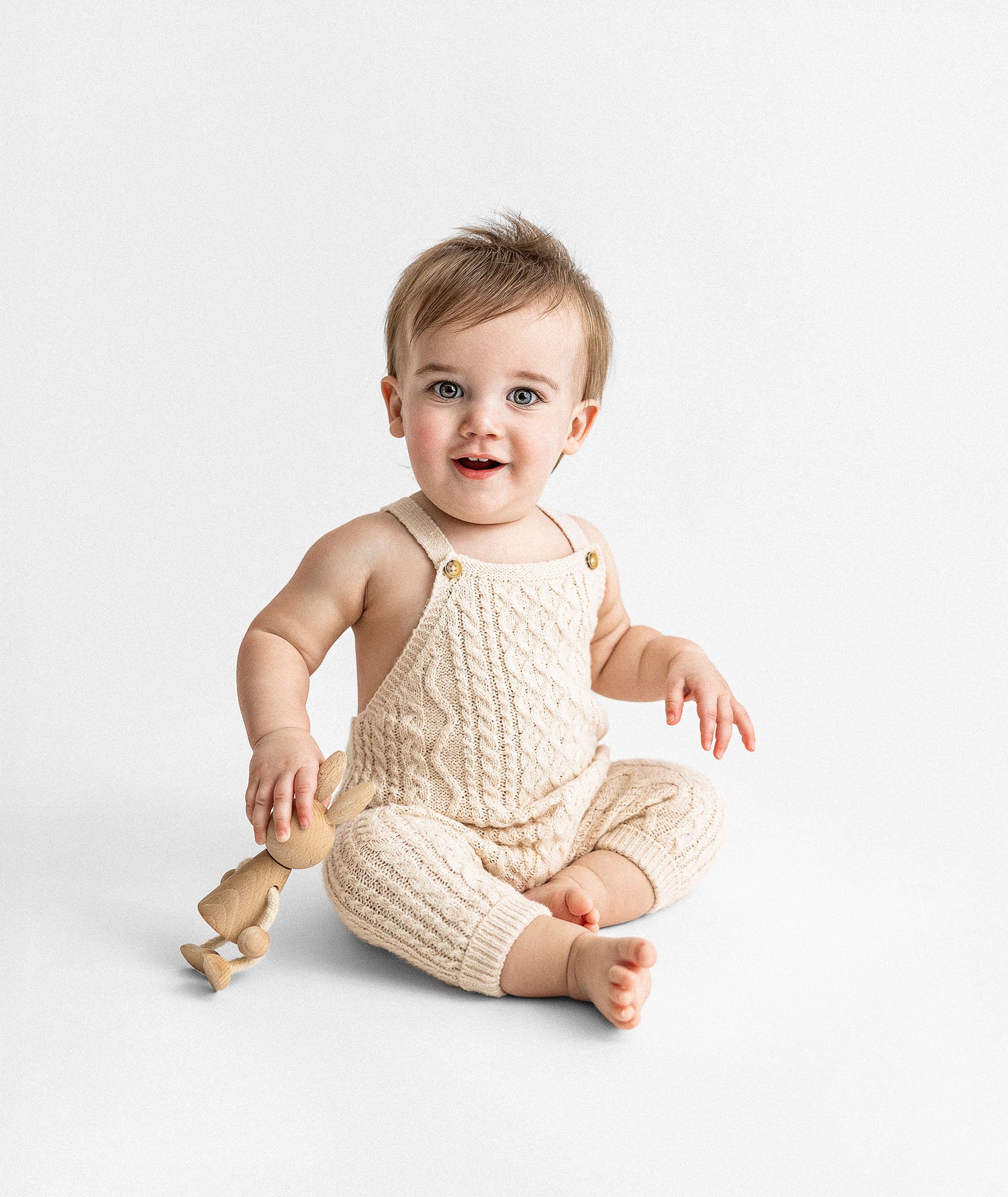 A baby in a cream knit outfit sitting on the floor holding a wooden toy bunny, with a white background.