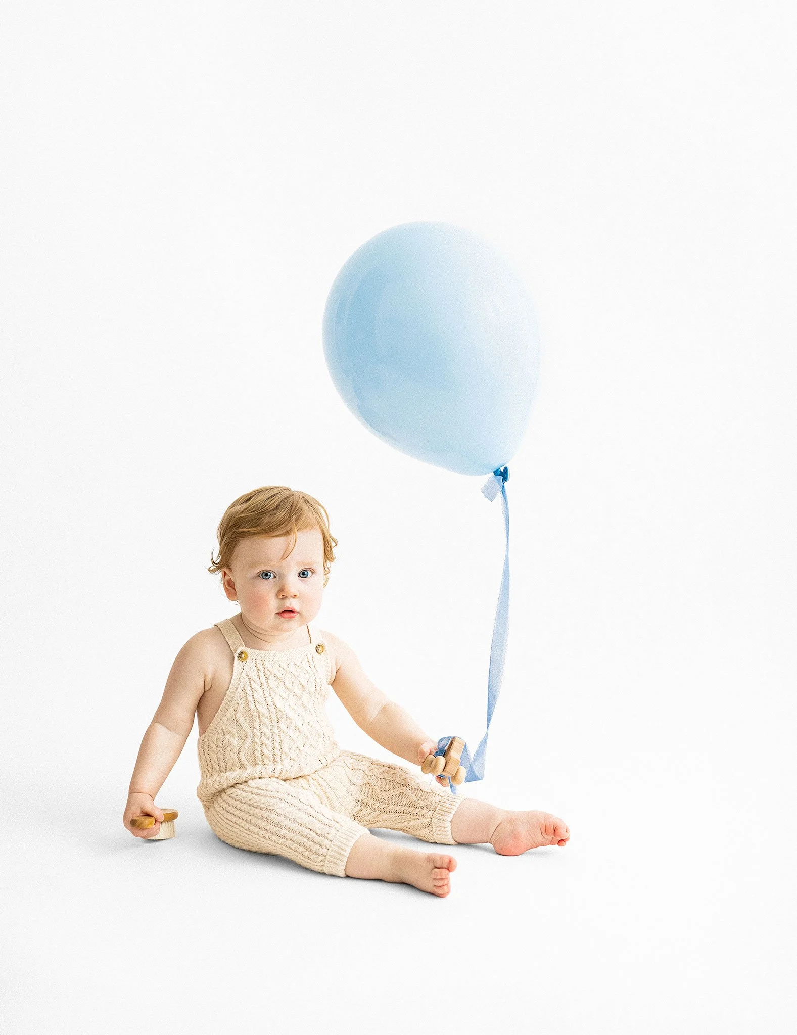 A young girl with light skin and blue eyes sitting on the floor, holding a wooden toy in one hand and a small skate in the other, dressed in a beige knitted outfit, with a light blue helium balloon floating above her.