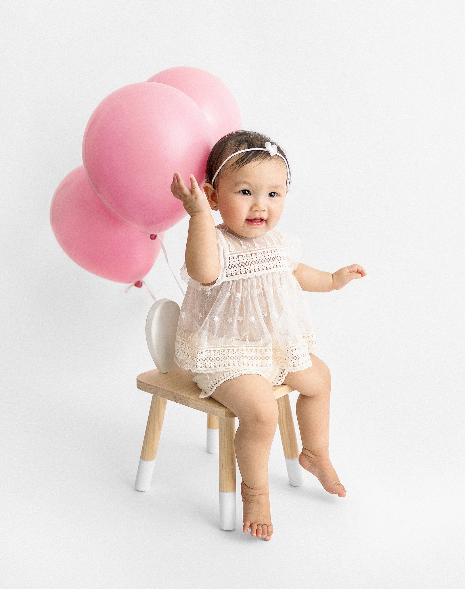 A baby in a white dress sitting on a small wooden chair, holding pink balloons.
