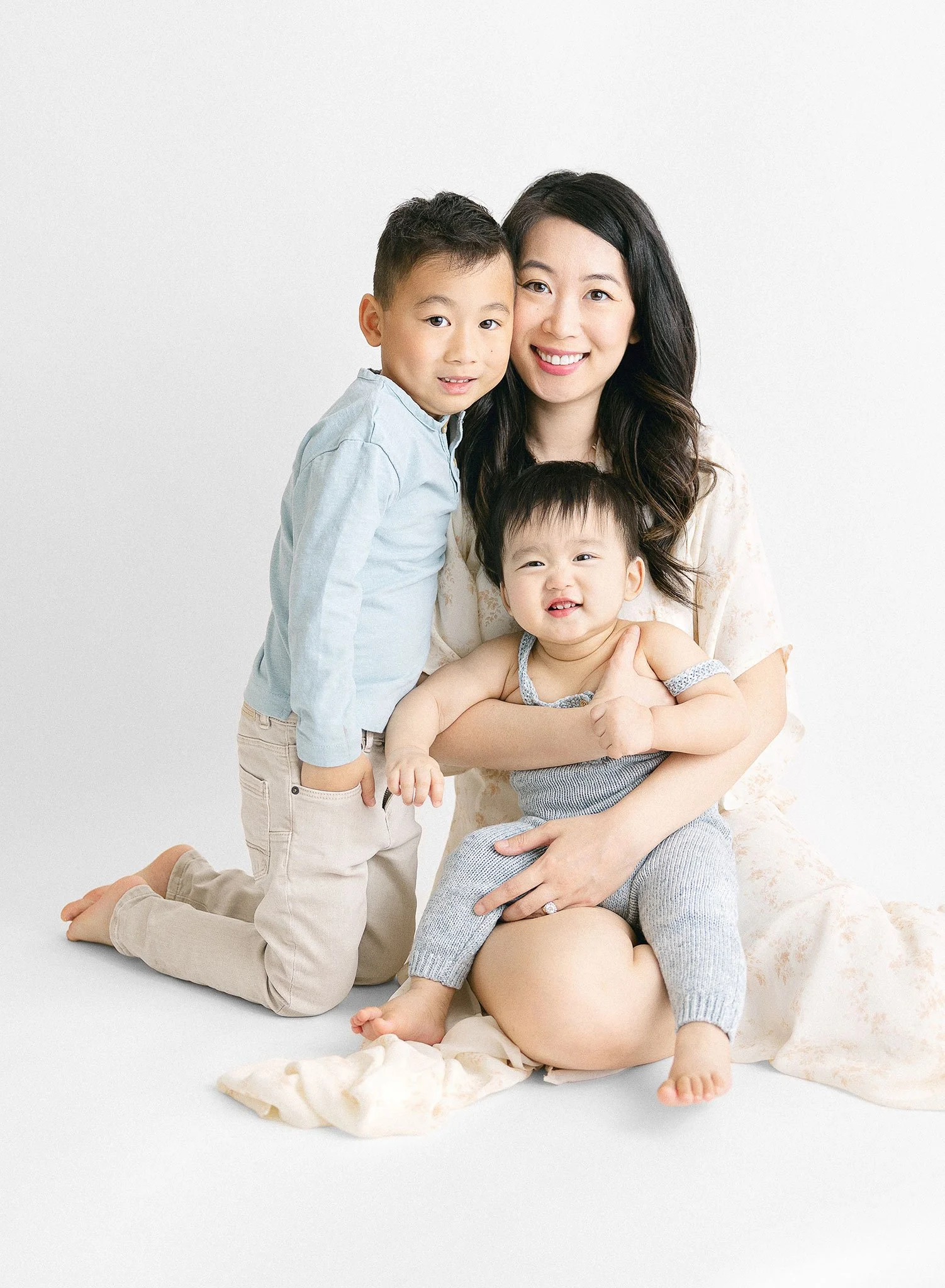 A woman is sitting on the floor, smiling, and holding a baby. A young child is leaning against her, also smiling. The setting is bright with a soft, light backdrop.