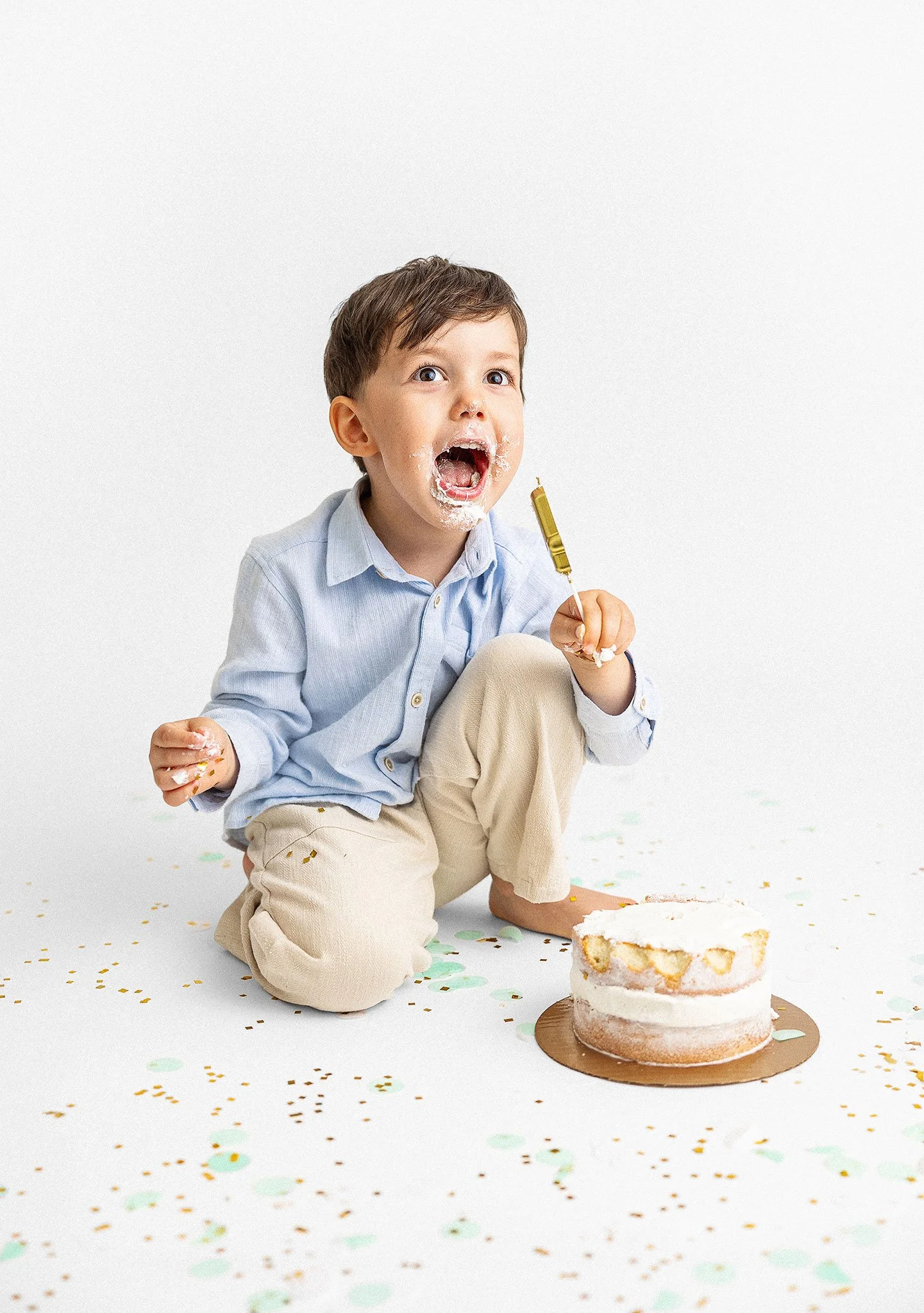 Child in blue shirt and beige pants with cake on their face, holding a cake spatula, sitting next to a partially eaten layered cake on a white floor with confetti.