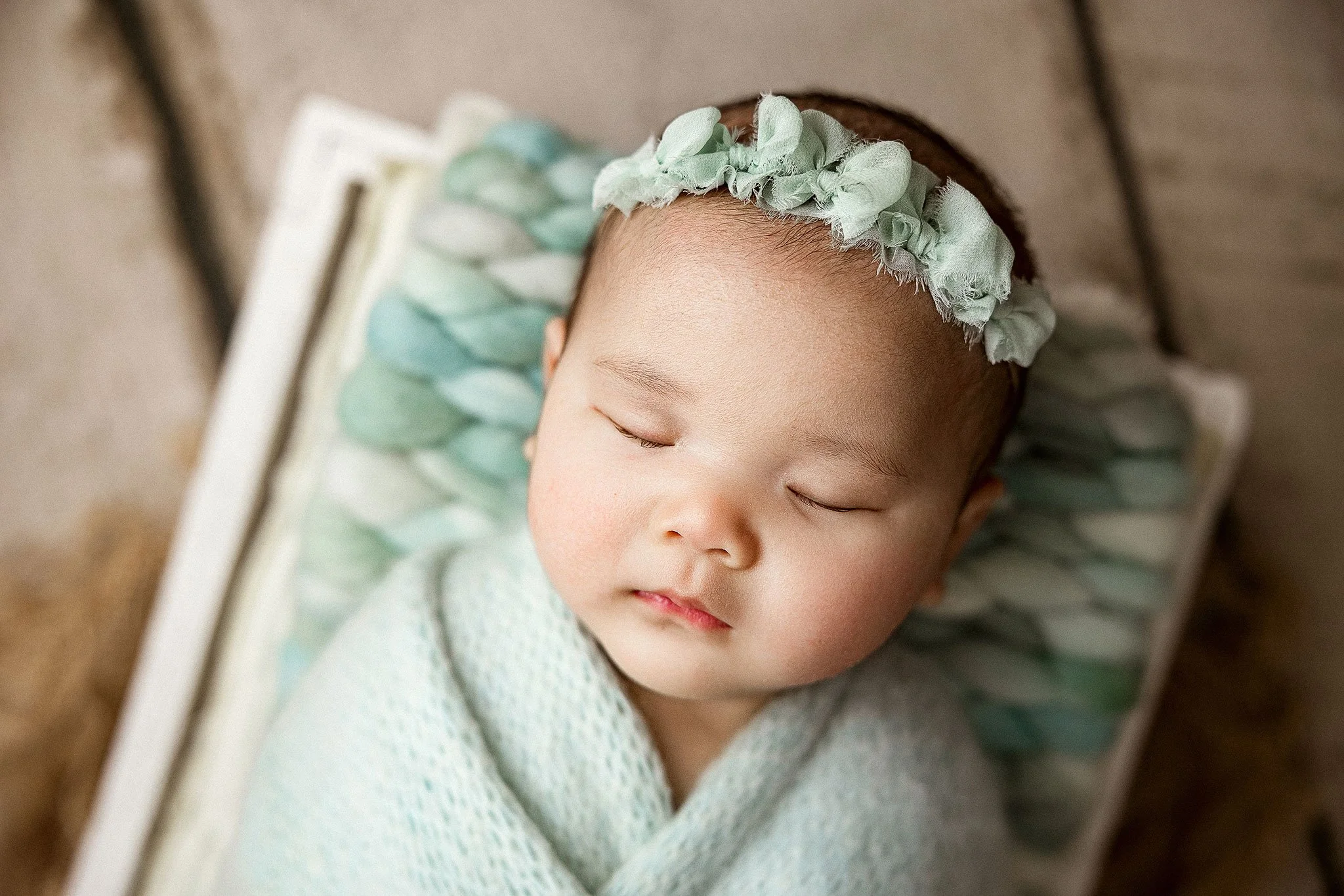 Close-up of a sleeping baby girl with a floral headband, wrapped in a light blanket, lying on a patterned pillow.