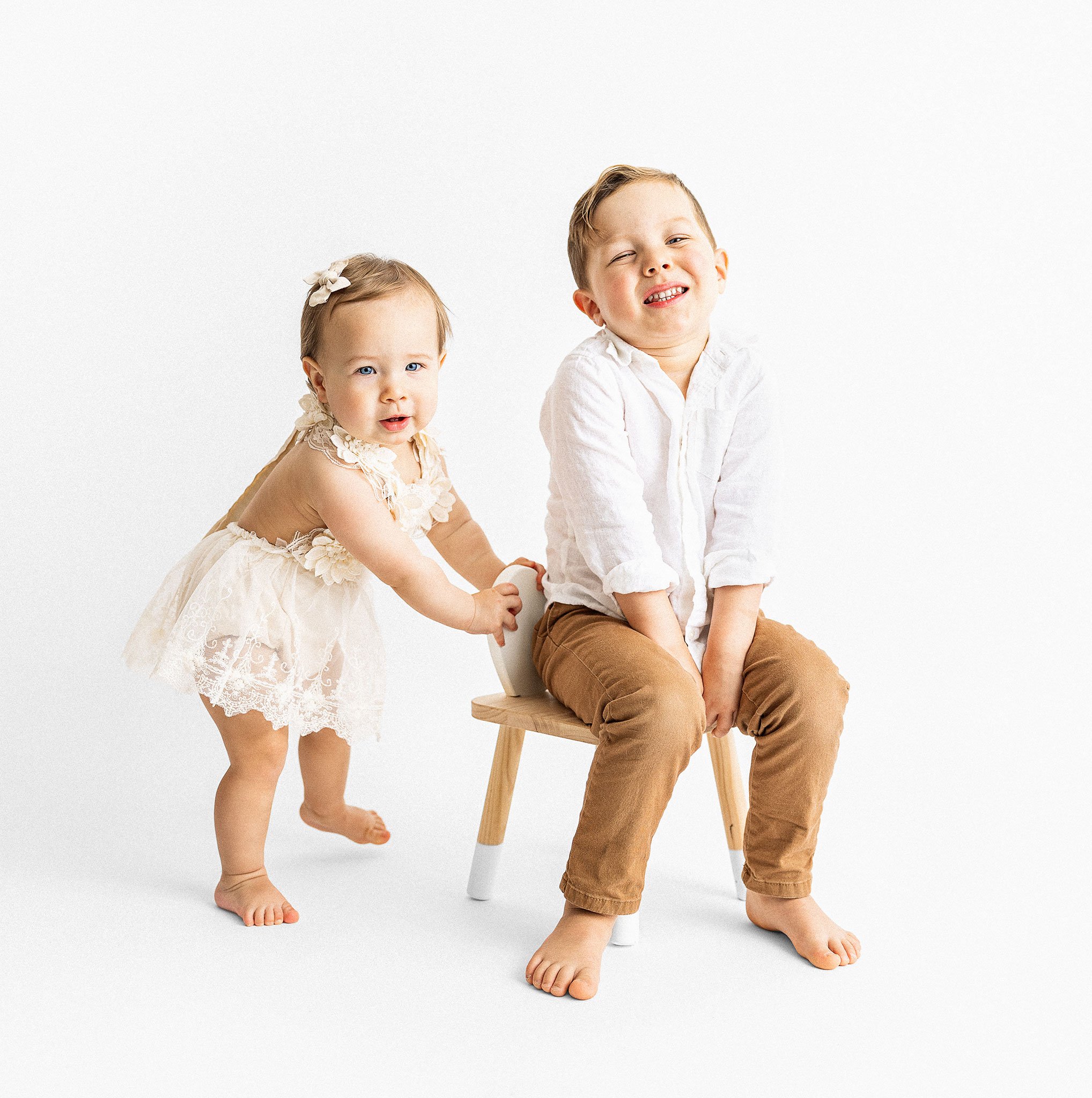Two young children in a bright studio setting; a girl wearing a lace dress stands while a boy in a white shirt and brown pants sits on a wooden chair, both smiling.