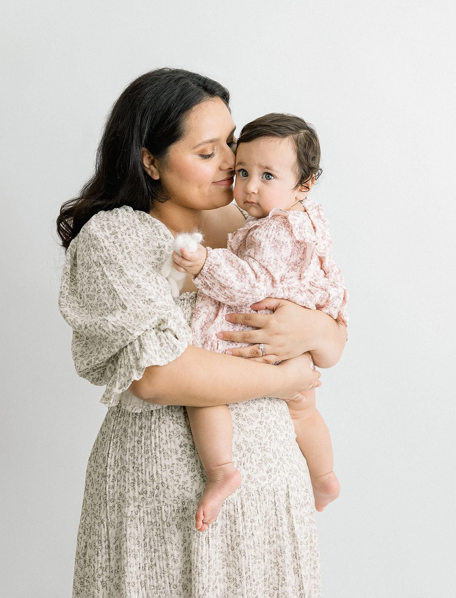 A woman gently holding a baby, both wearing light-colored outfits. The woman is kissing the baby's cheek while the baby looks at the camera. The background is plain and white.