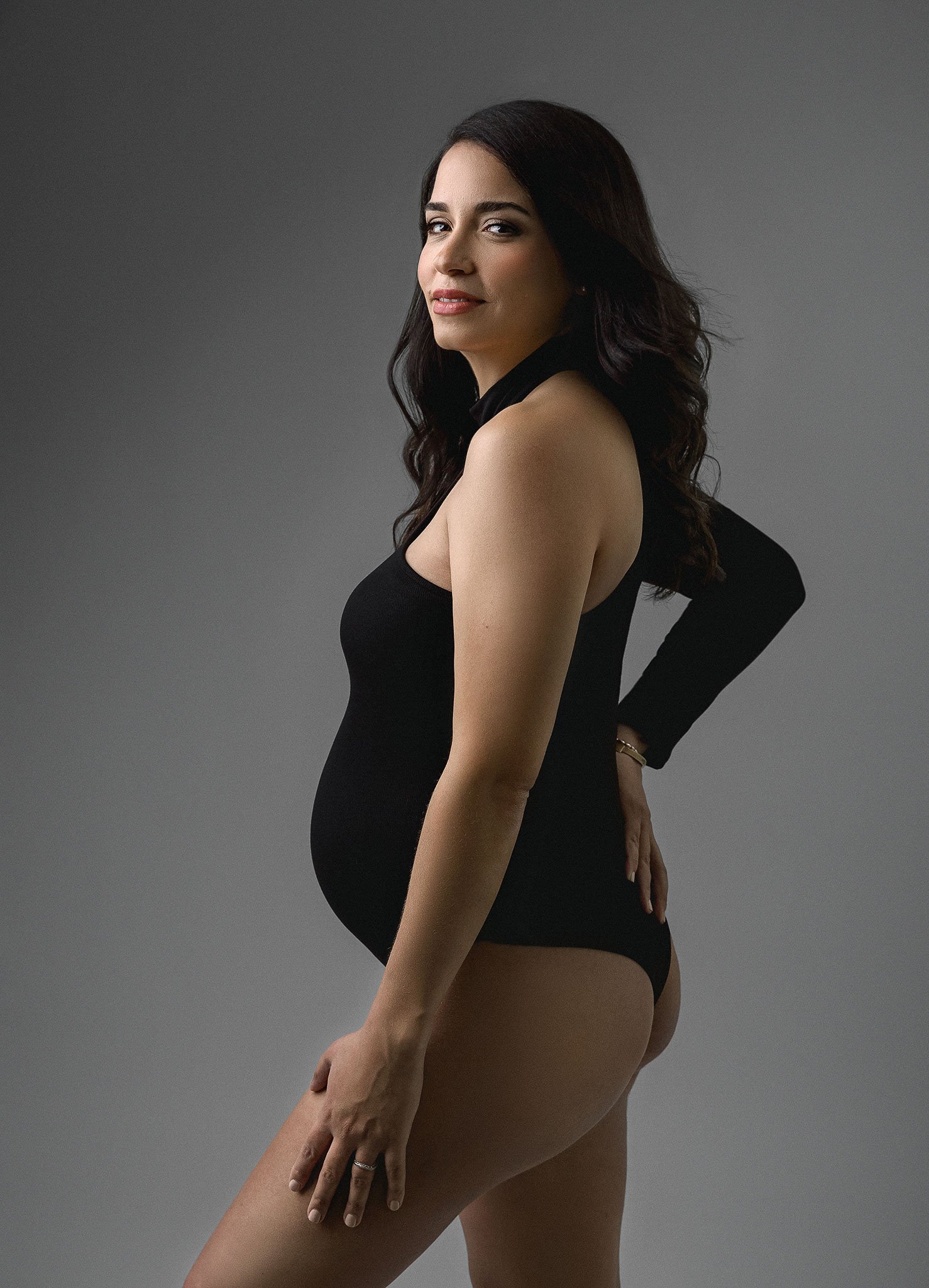 Pregnant woman in black bodysuit, side profile, hand on hip, against gray background.