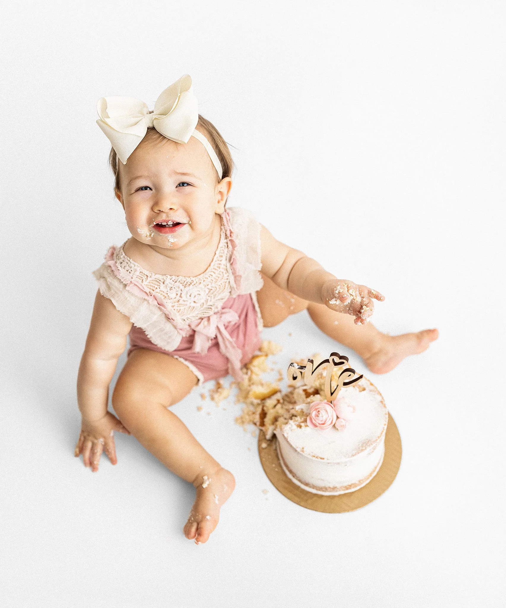 A young girl with a large cream-colored bow headband, sitting at a table with a birthday cake decorated with pink roses and a cake topper that says 'love.' The girl is smiling with some cake on her face and hands.
