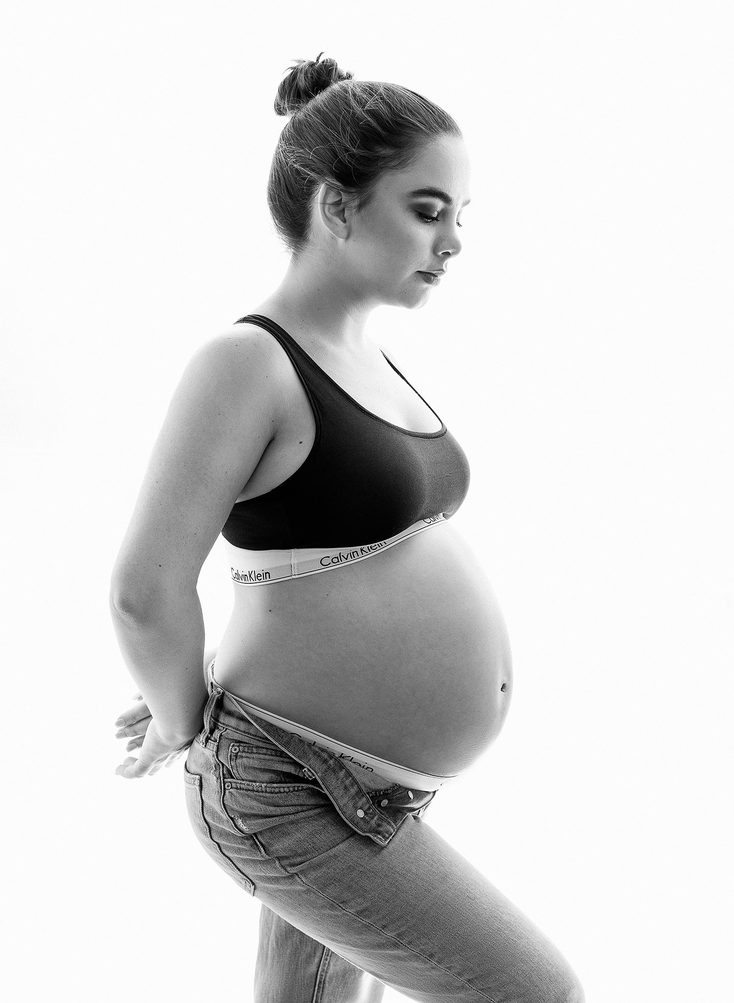Pregnant woman in profile wearing Calvin Klein sports bra and jeans.