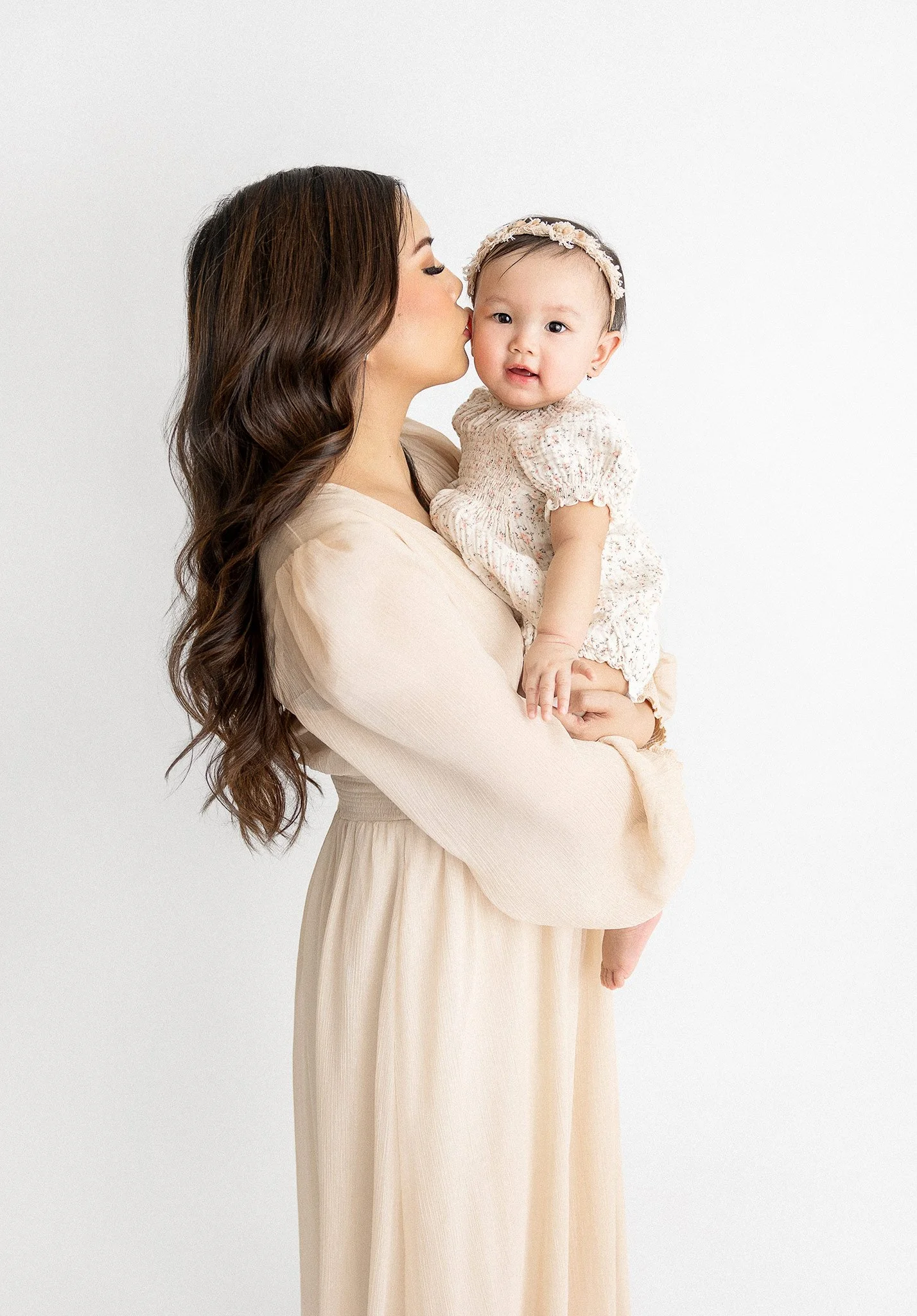 Woman with long wavy brown hair holding a baby girl in a cream dress and floral headband, woman is kissing baby's temple against a plain white background.