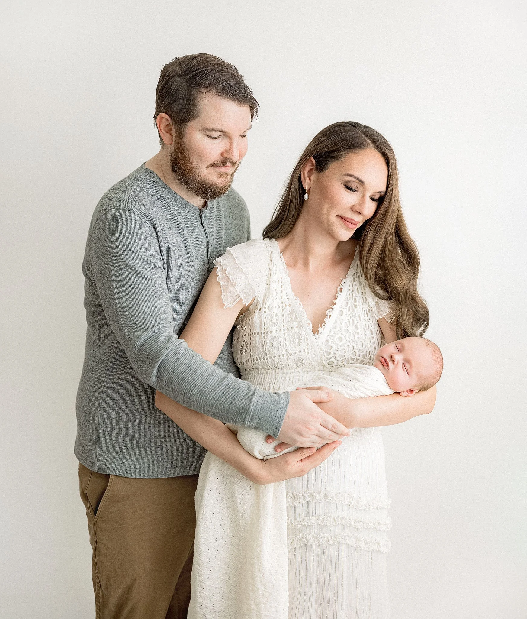 A couple holding a newborn baby, the woman is pregnant, standing against a plain light-colored background.