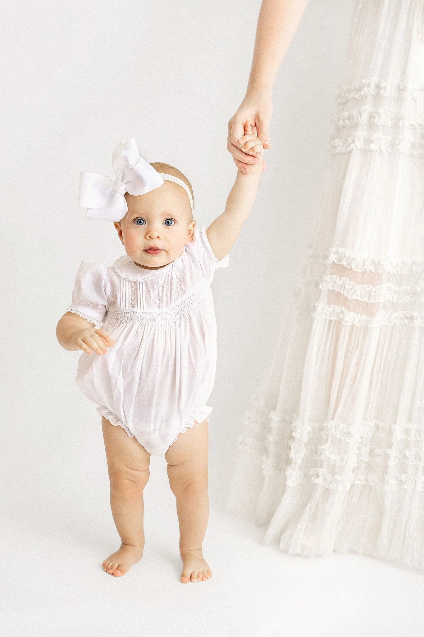 A baby in a white outfit with a large white bow headband is holding the hand of an adult wearing a white dress.
