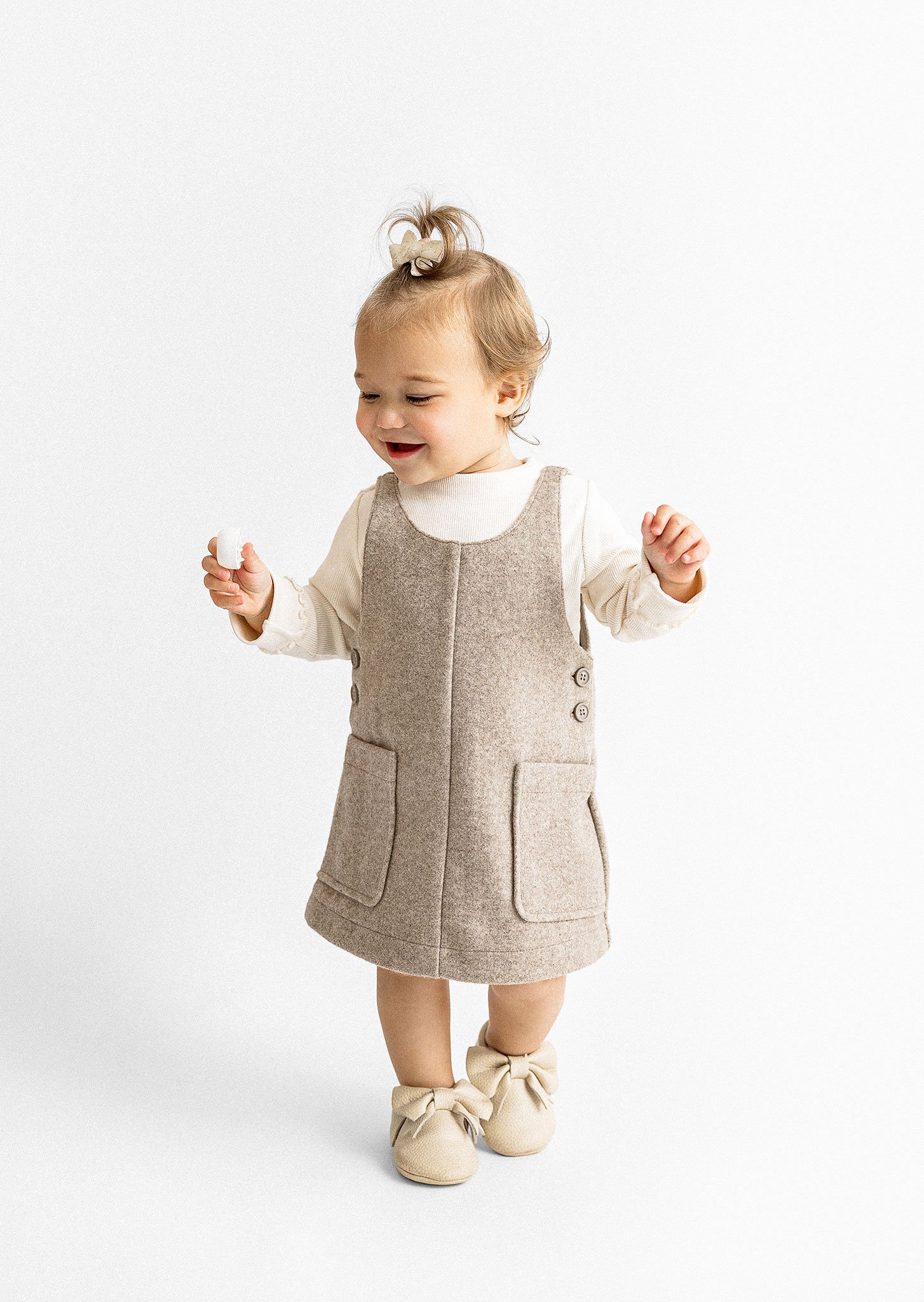 A toddler in a beige dress and white top, with beige shoes, smiling and holding a small item.