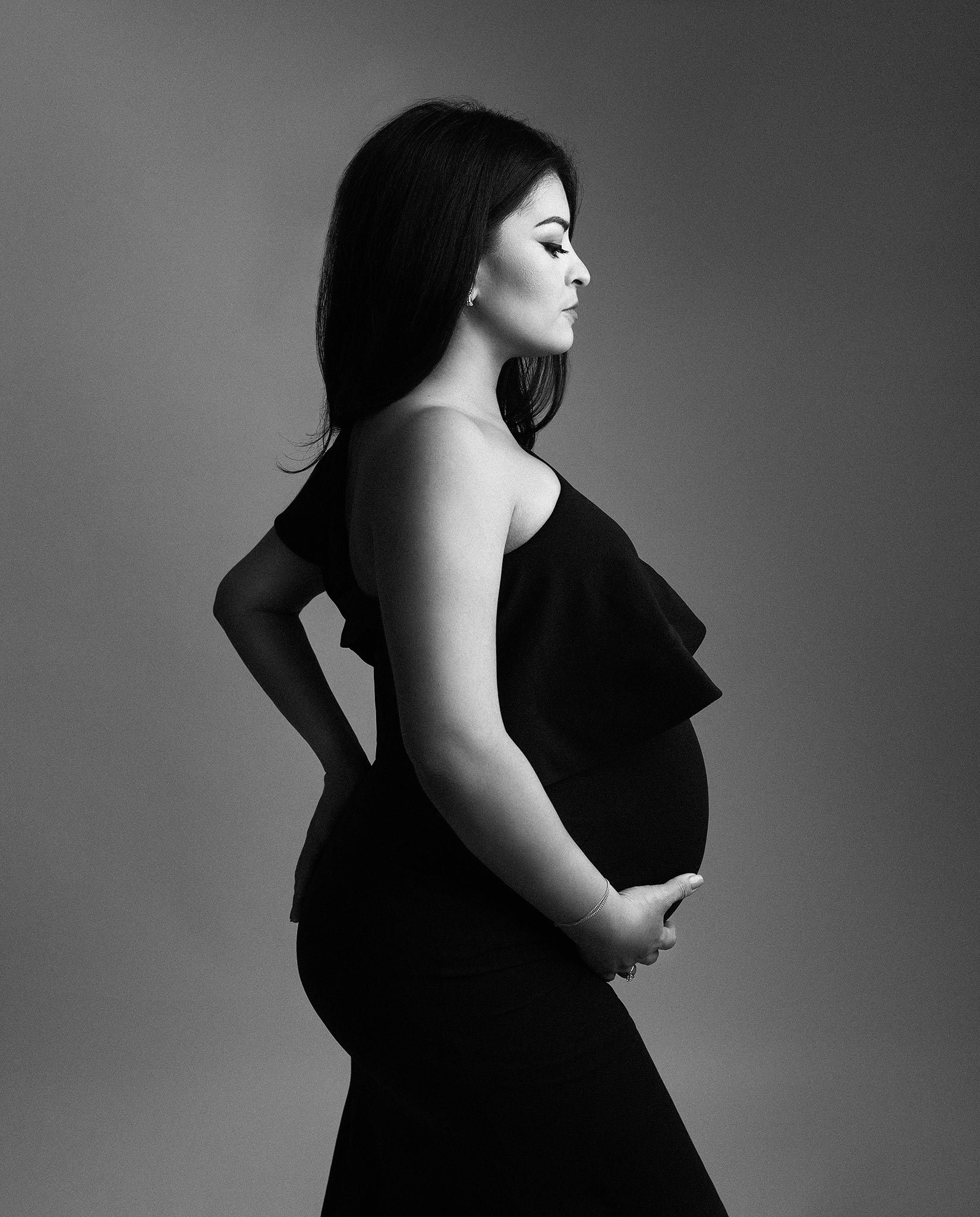 Black and white profile portrait of a pregnant woman wearing a black dress, holding her belly, standing against a plain background.