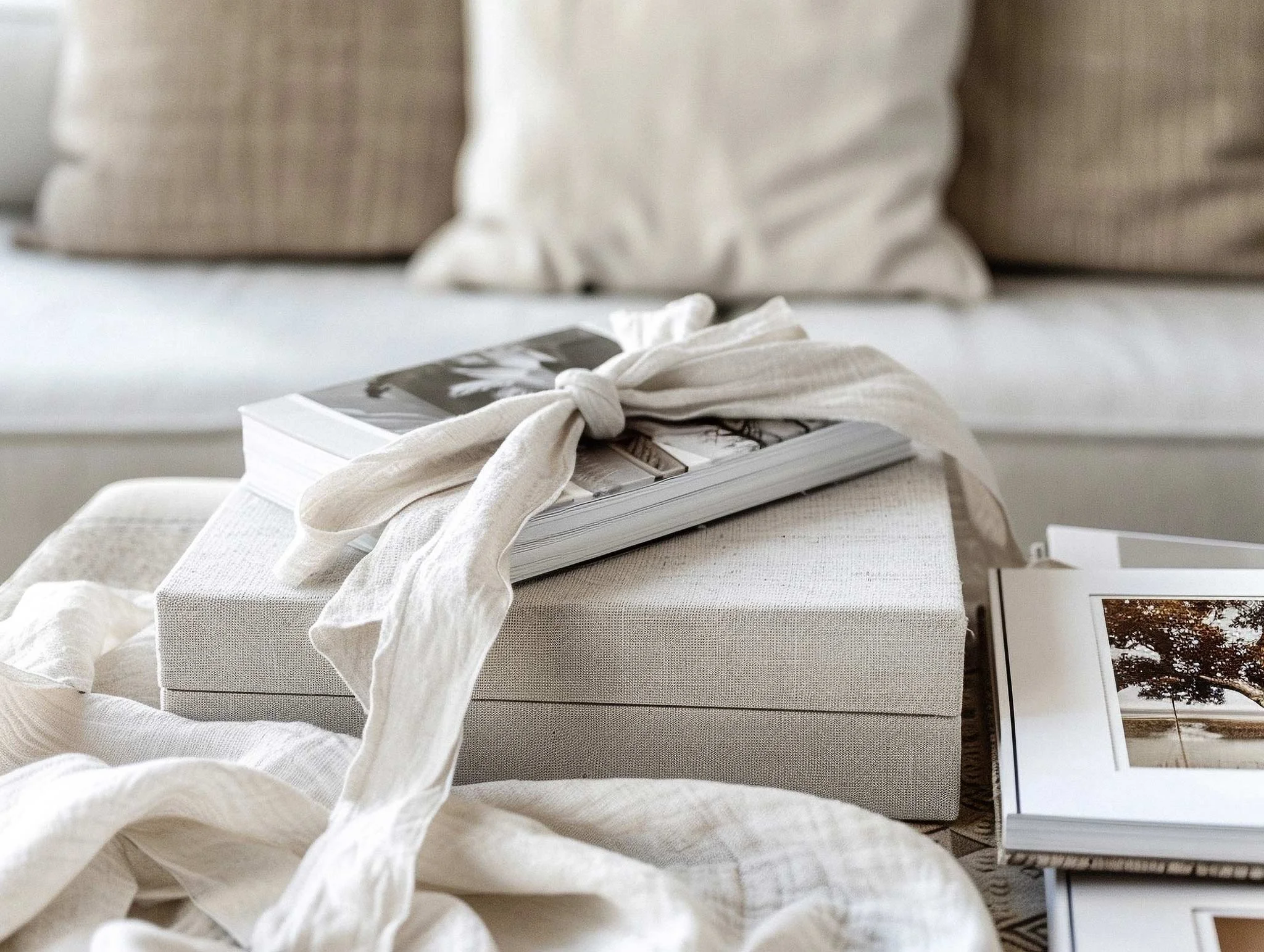 A stack of books with a cloth napkin on top, placed on a table with a textured fabric cover, in a cozy room with cushions and framed artwork on the wall.