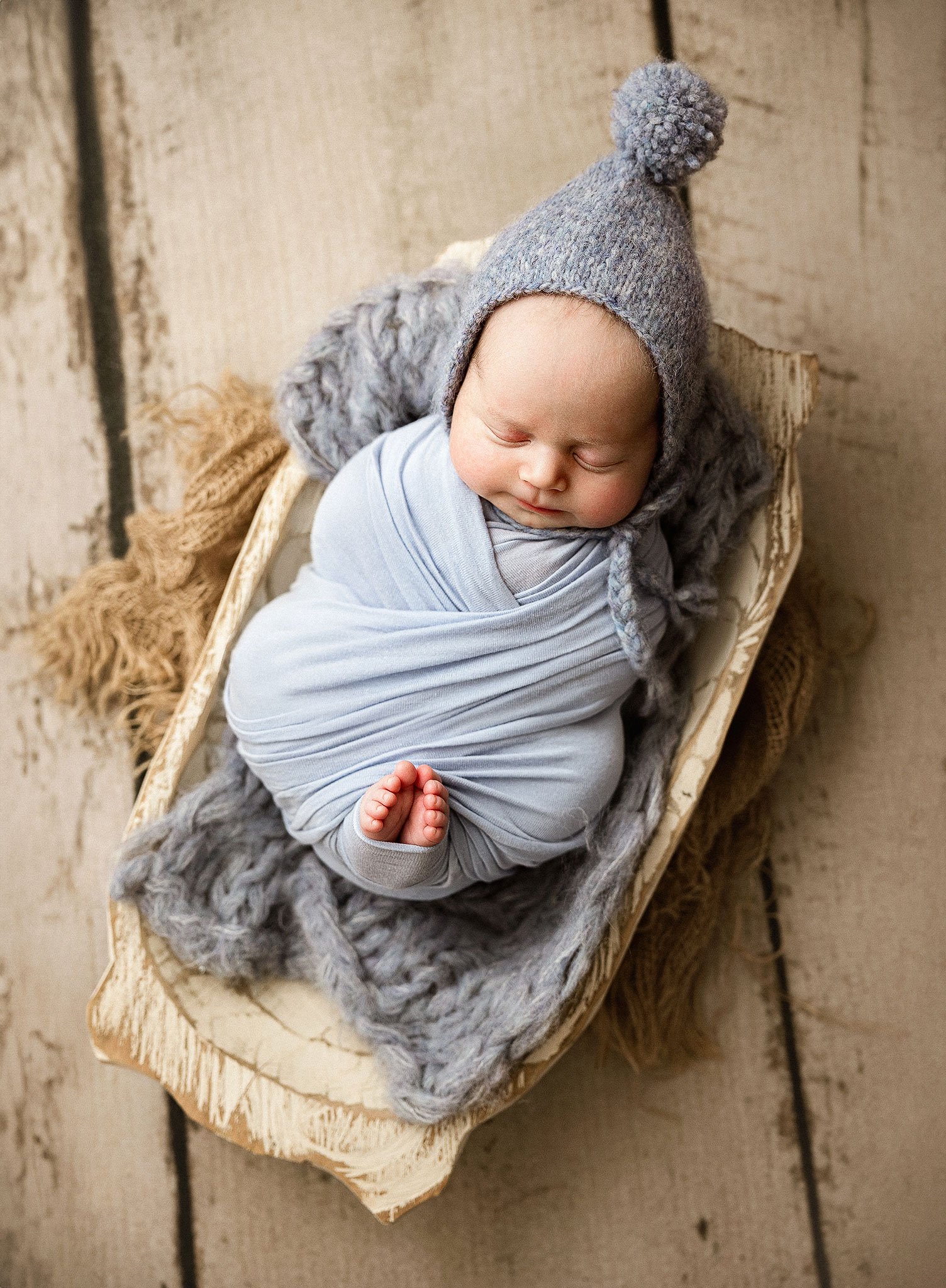 Newborn baby swaddled in blue blanket, wearing a knitted gray hat, sleeping in a rustic wooden bowl on a textured wooden surface.