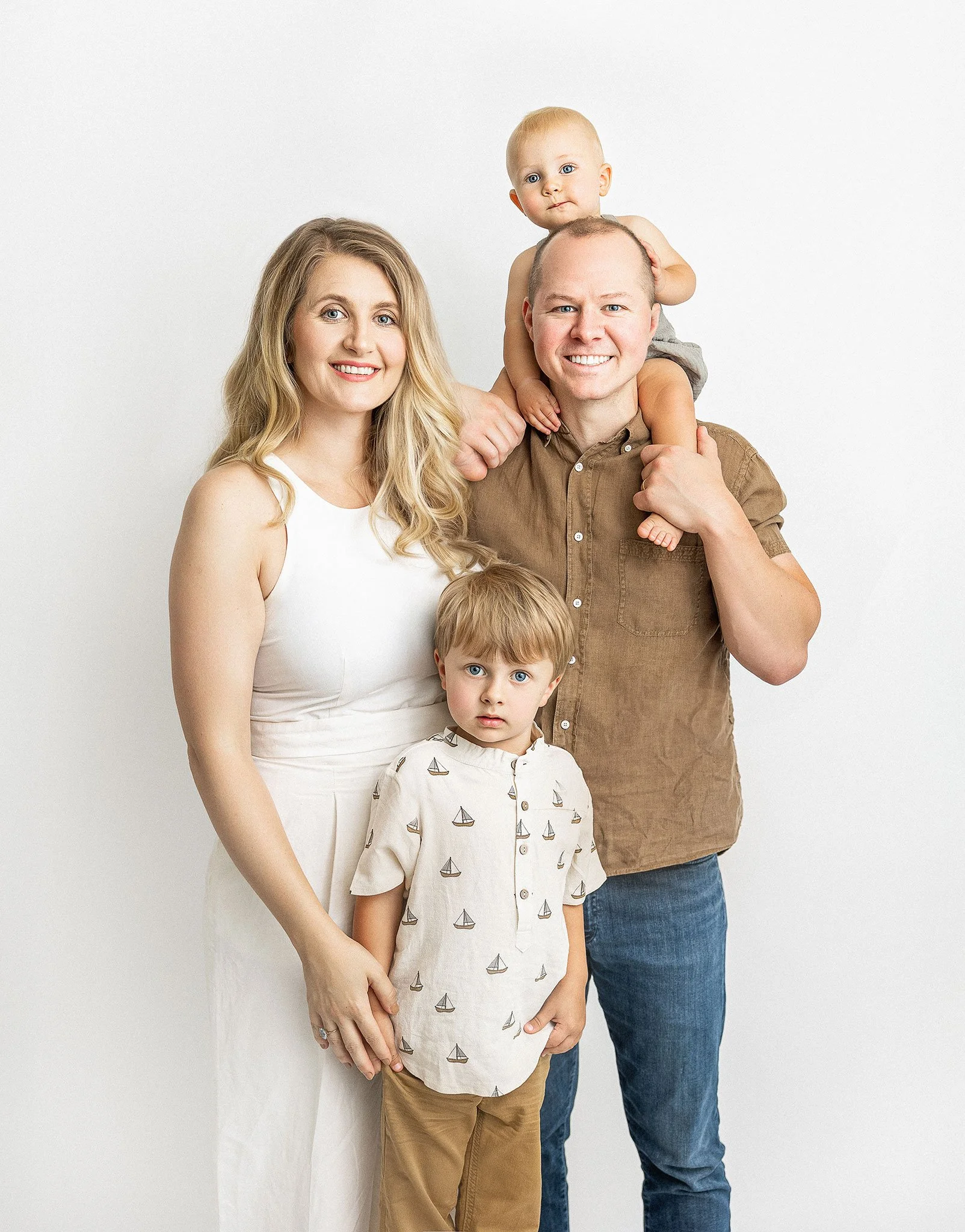 A smiling family with two parents, a young child, and a baby. The woman is wearing a white dress, and the man is dressed in a brown shirt and jeans. The young child stands in front, wearing a shirt with boat patterns. The baby is seated on the man's 