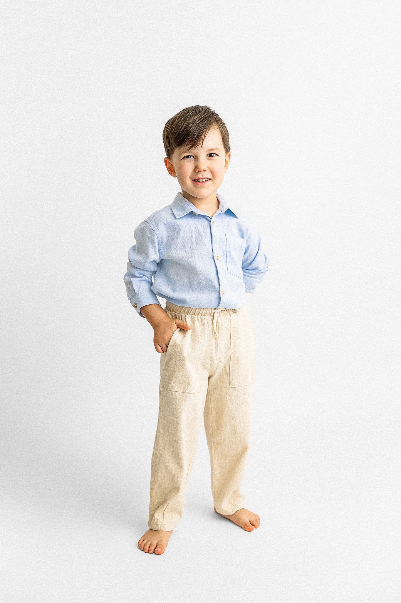 Young boy smiling, wearing a light blue button-up shirt and beige pants, with hands in pockets.