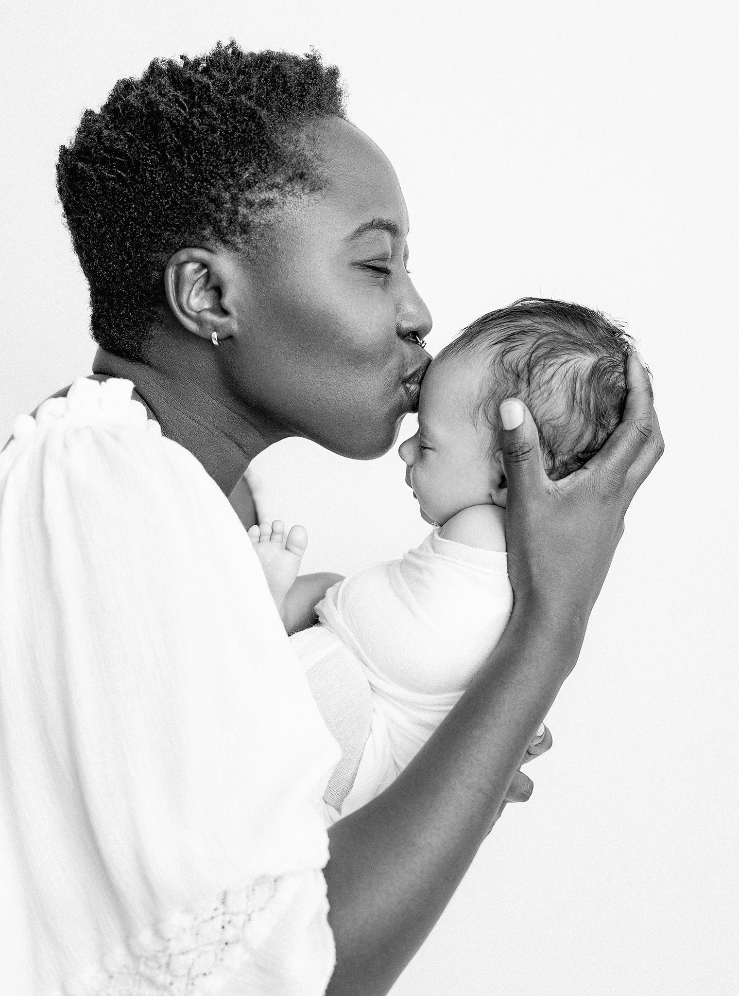 A woman with braided hair gently kissing the forehead of a sleeping baby girl with wet hair, in black and white.