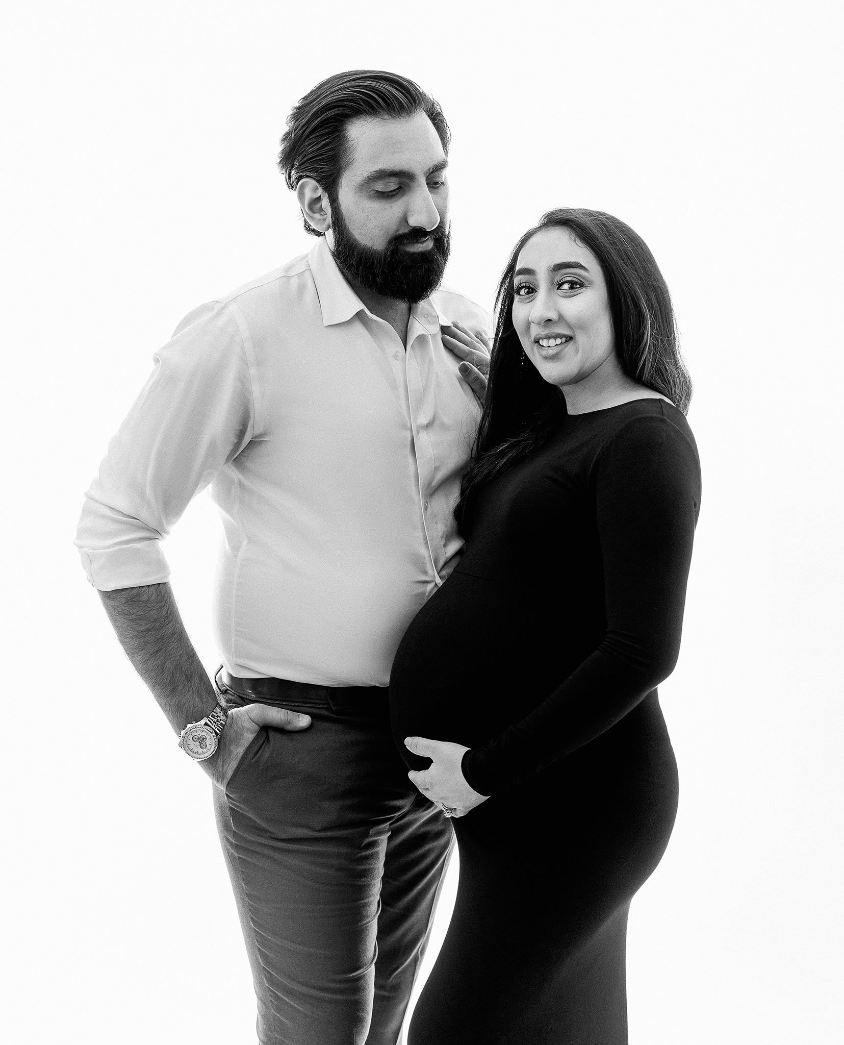 A black and white photo of a man and a pregnant woman, standing close with the man's hand on the woman's shoulder. The woman is wearing a form-fitting dress, holding her belly.