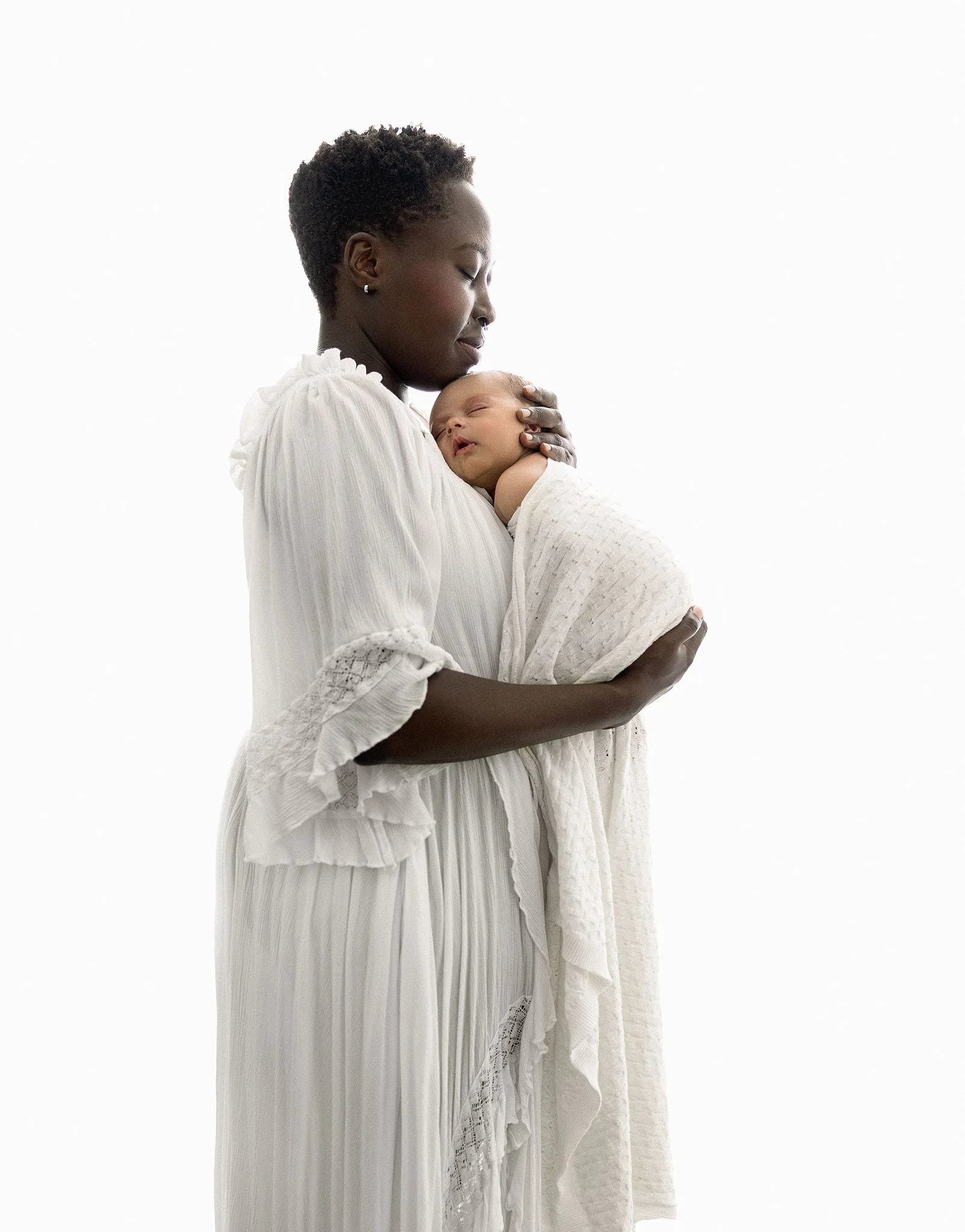 A woman with short curly hair and earrings holding a baby wrapped in a white blanket, both wearing white clothing, against a plain white background.