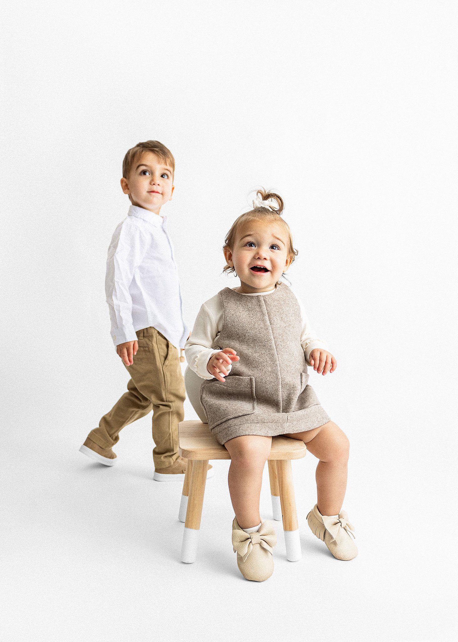Two young children in stylish clothing, one sitting on a stool and one standing, with a white background.
