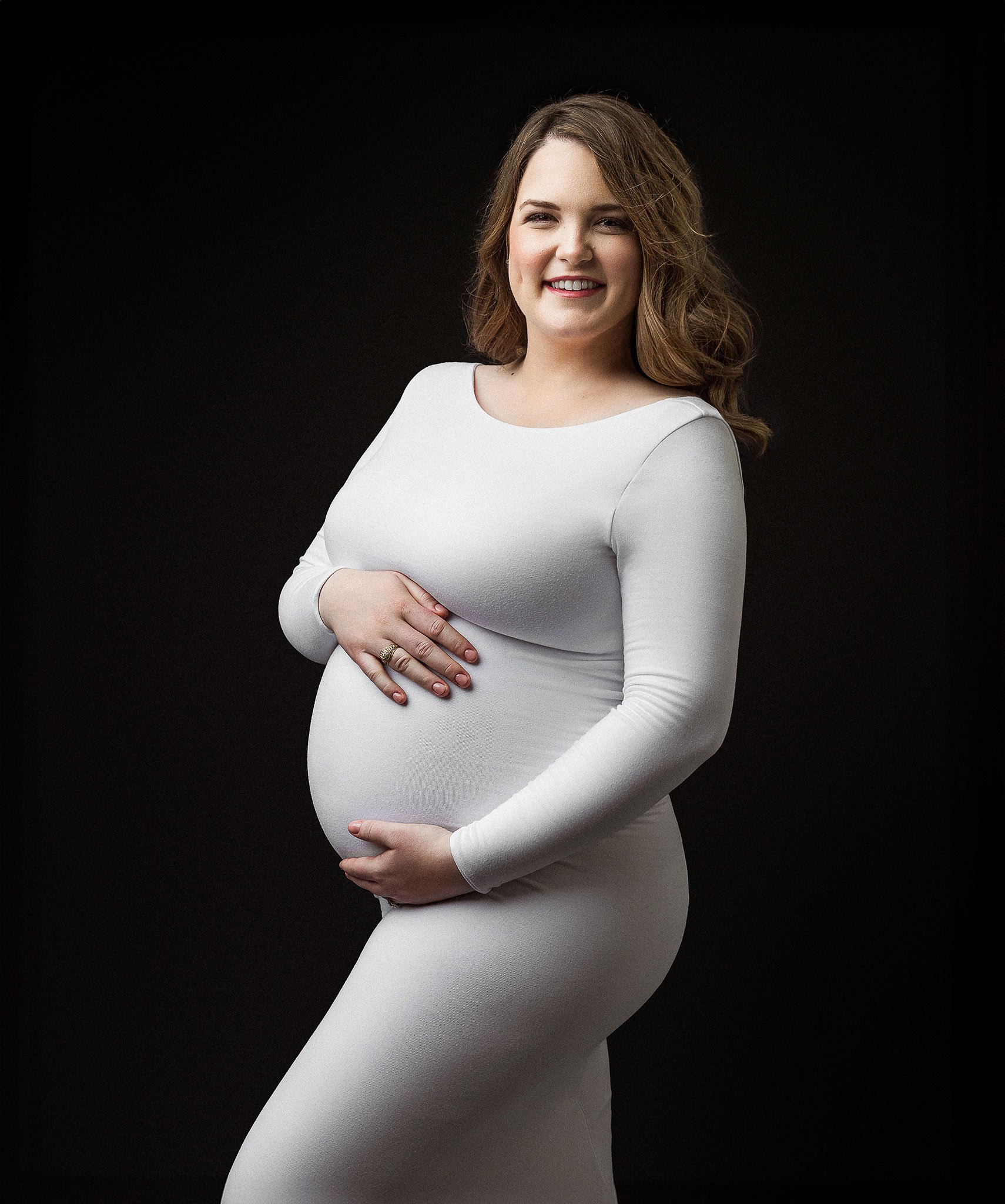 Pregnant woman in white dress holding her belly, smiling, with a dark background.