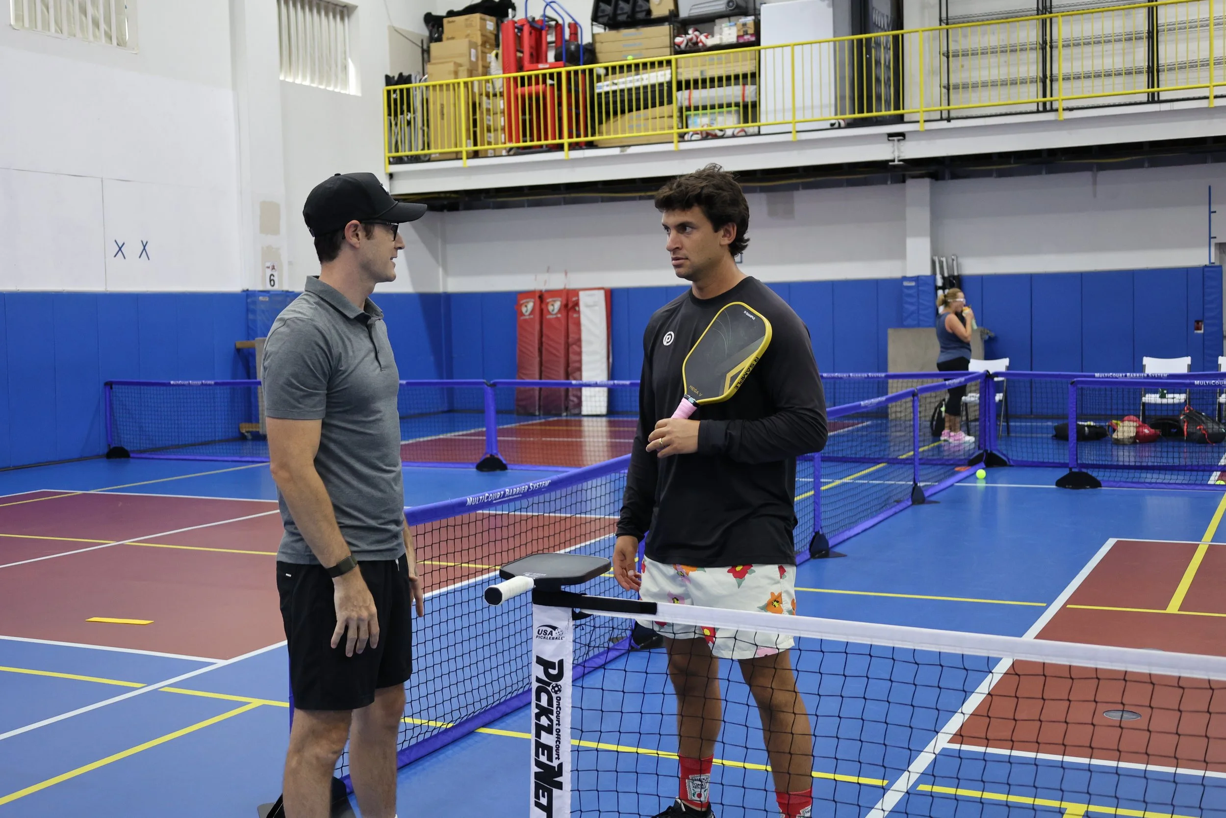 Two men having a conversation near a pickleball net in an indoor gym. One man wears a gray shirt and a black cap, and the other man wears a black long sleeve shirt and colorful shorts, holding a pickleball paddle. In the background, a woman is standing near the wall, talking on her phone with equipment and a yellow railing above.
