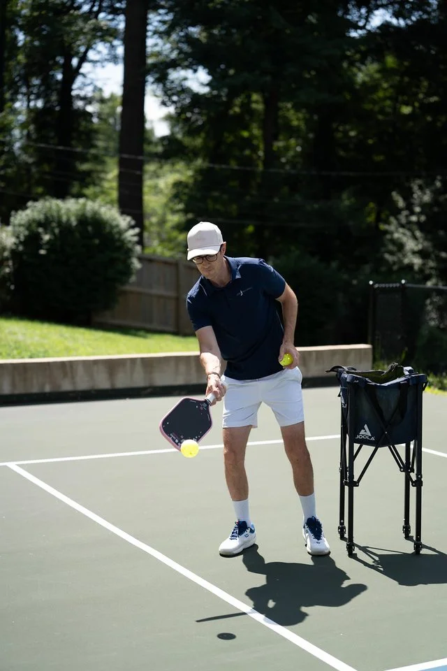 A young man playing pickleball on an outdoor court, holding a pickleball paddle and a tennis ball, with a ball cart nearby.