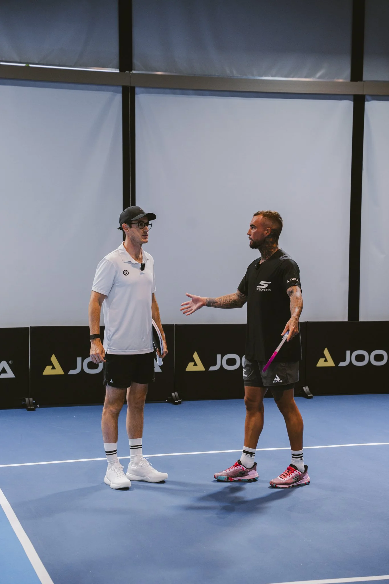 Two men standing on an indoor tennis court, engaged in conversation. One man is wearing a white polo shirt, black shorts, white sneakers, and a black cap. The other man is wearing a black sports shirt, black shorts, pink sneakers, and has tattoos on his left arm. They appear to be coaches or players, with a tennis racket in hand of the man in black.
