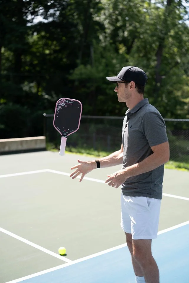 A man in a gray polo shirt, white shorts, and a baseball cap appears to be juggling tennis balls on an outdoor tennis court, with a tennis racket floating in mid-air nearby.