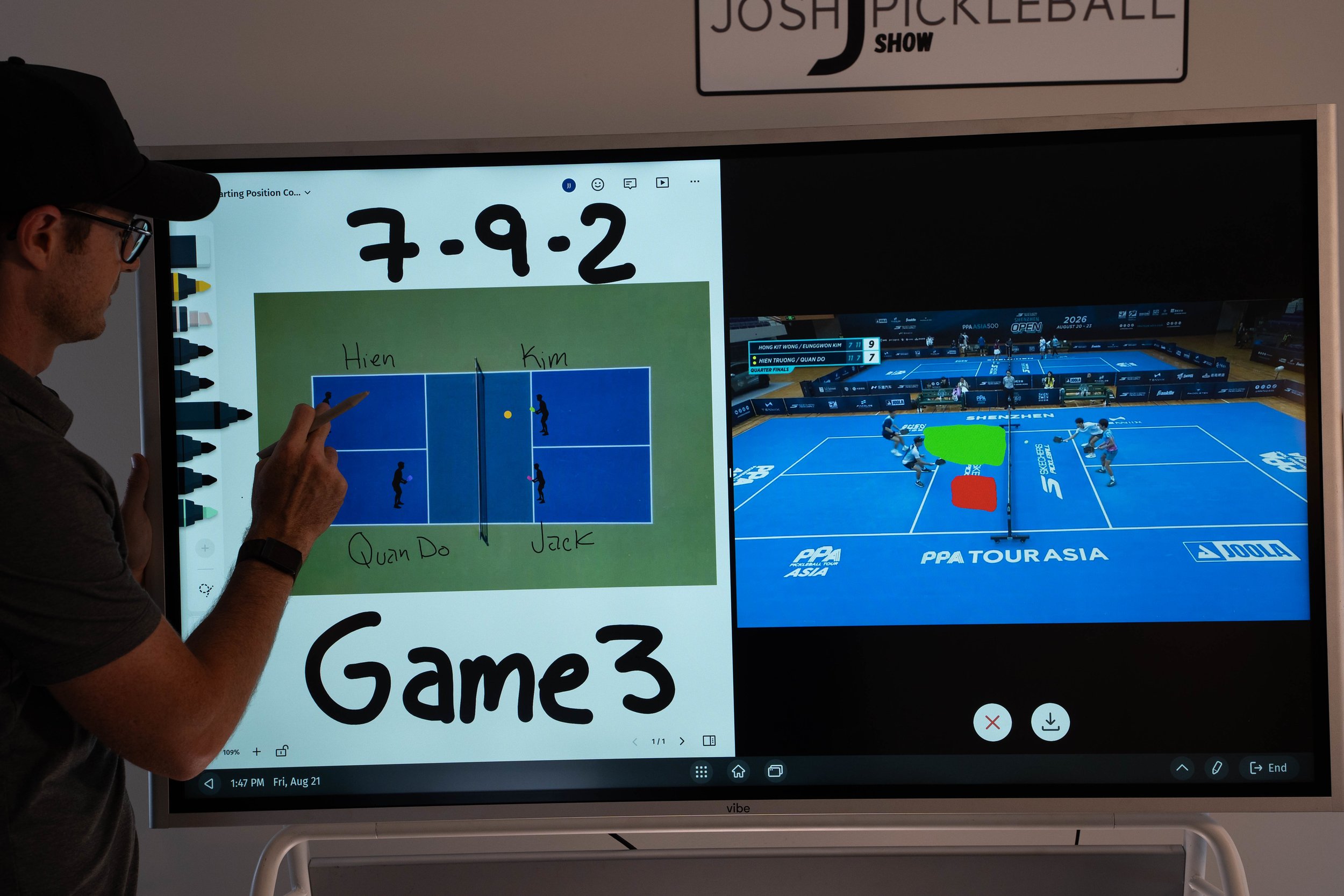 A man is pointing at a digital screen with handwritten notes and drawings, including a math problem and some stick-figure illustrations. Behind him, a large monitor displays a badminton court with players, score, and event information.