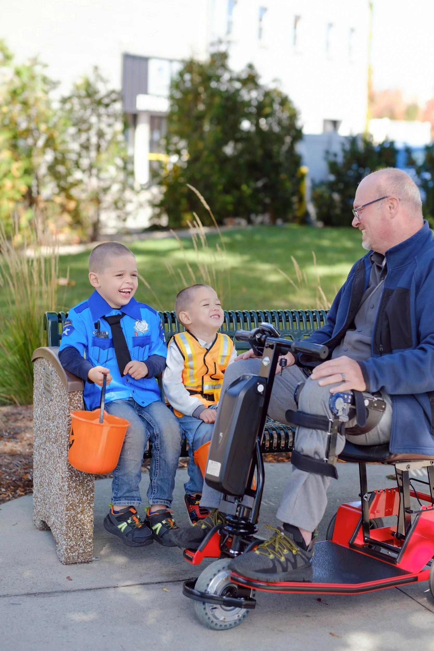 Two smiling children sitting on a park bench with a man riding a mobility scooter on a sunny day.