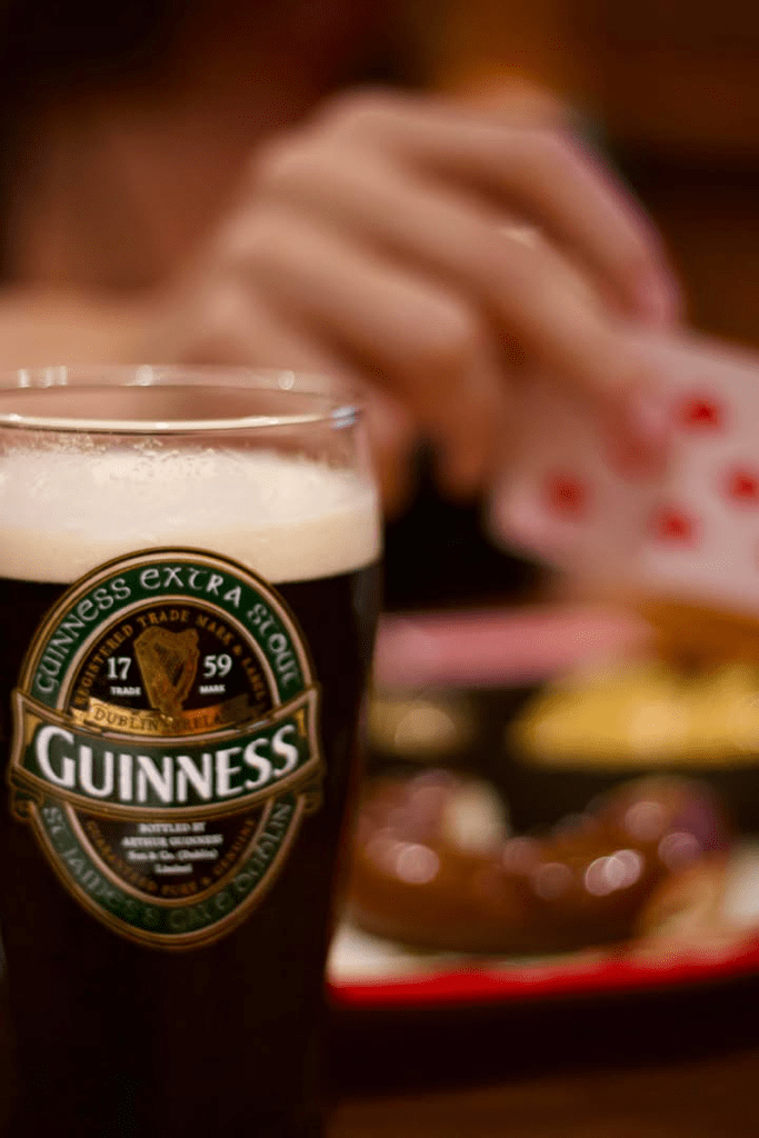 A pint glass of Guinness beer with foam on top, with a hand holding playing cards in the background.
