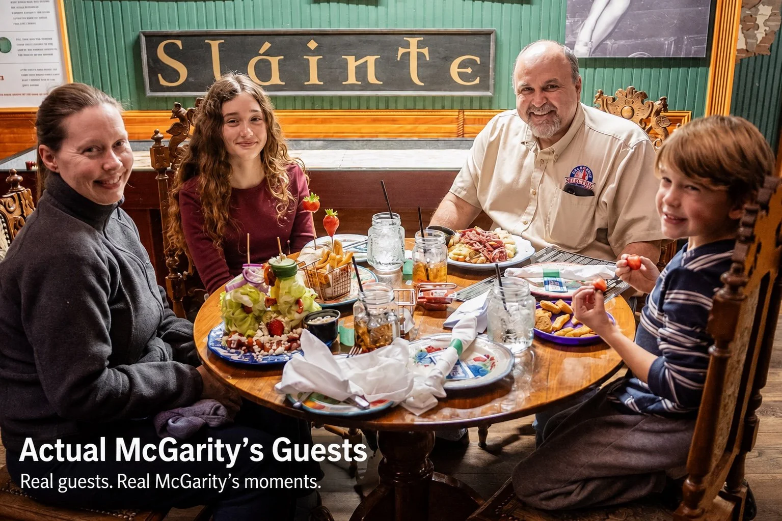 Four people sitting around a wooden table with food and drinks, smiling at the camera in a restaurant with a sign that says 'Sláinte' on the wall.